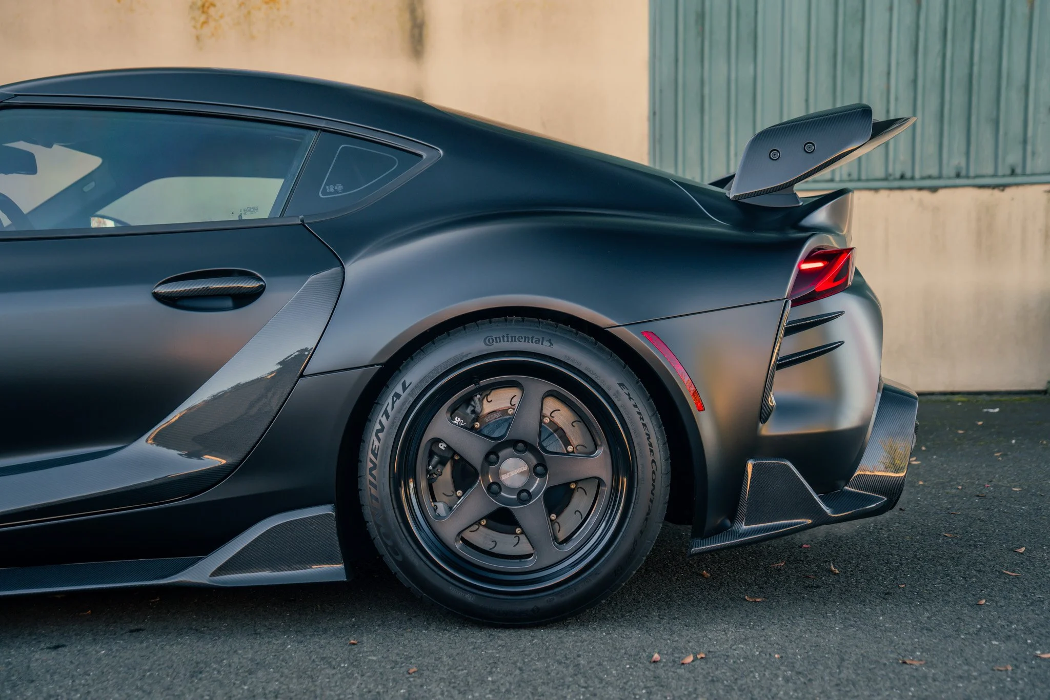 Close-up of the rear side of a sleek, black sports car with a large rear spoiler, aerodynamic vents, and Michelin tires parked on dark pavement.