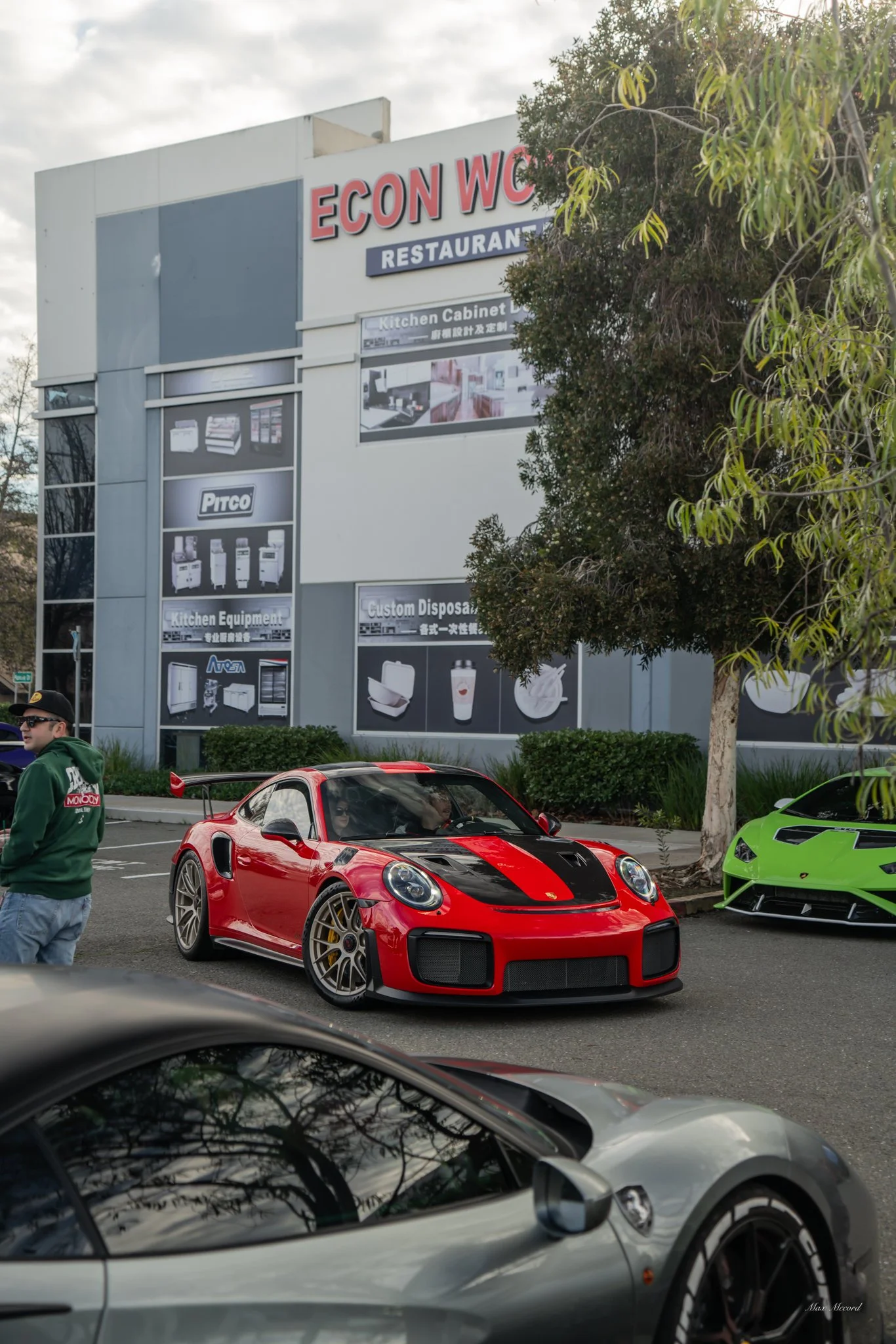 A red sports car with black racing stripes parked in a lot next to a green Lamborghini. A person in a green hoodie and cap is walking nearby. A building with a sign reading 'ECON WC RESTAURANT' and advertisements for kitchen equipment is visible in t