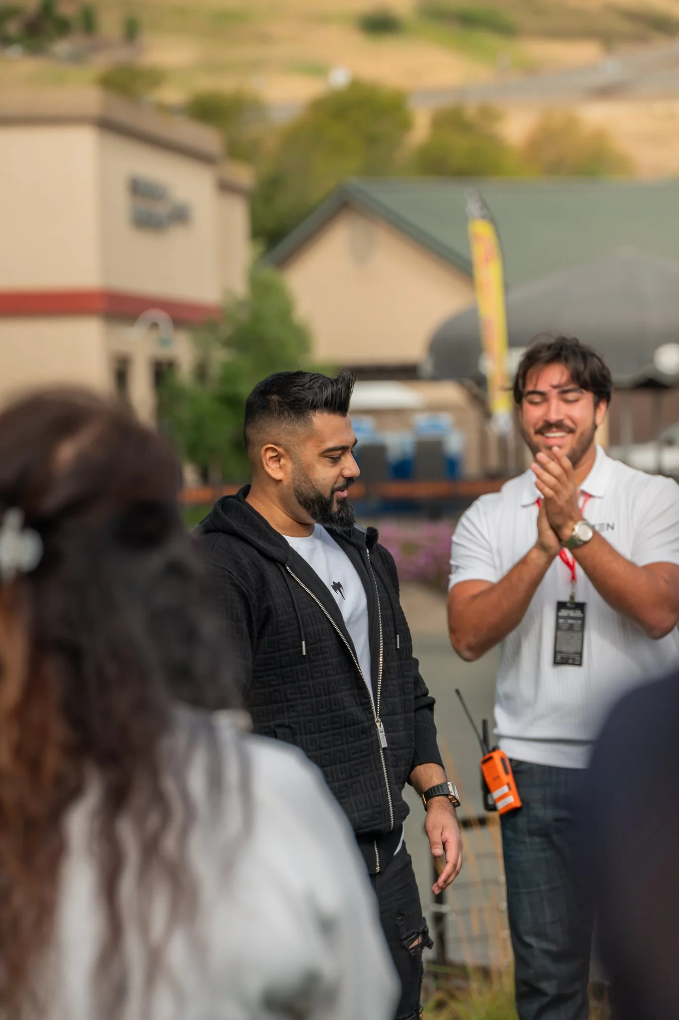 Two men standing outdoors, one with a black jacket and the other with a white polo shirt, engaged in a conversation or presentation, with blurred people in the foreground and a building and greenery in the background.