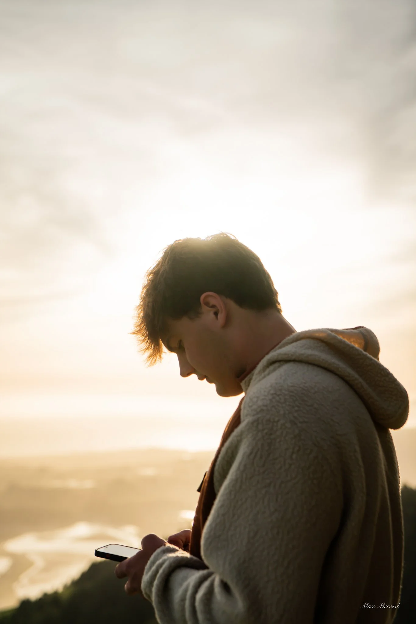 A young man with dark hair wearing a beige hoodie is looking down at his phone outdoors during sunset, with the sun behind him creating a glow around his head.
