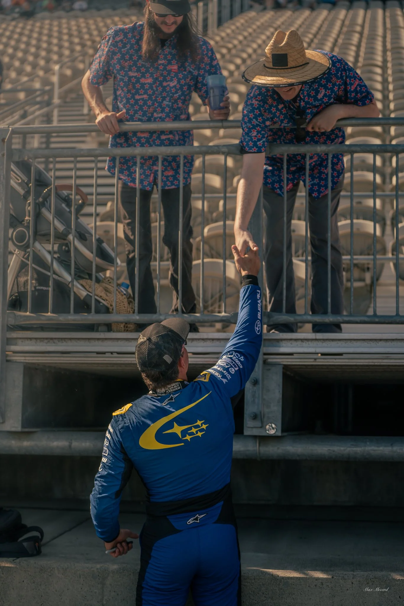 A race car driver in a blue racing suit is reaching up over a barrier to shake hands with two fans, one of whom is wearing a straw hat, at a racetrack with empty stadium seats in the background.