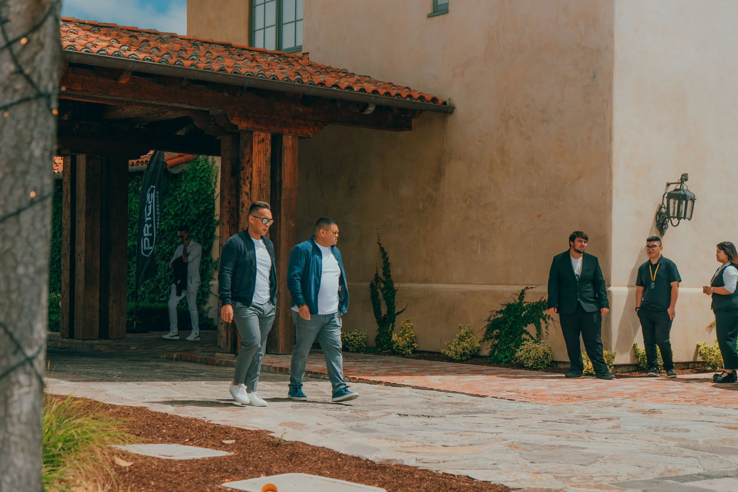 Group of people walking and standing outside a building with stucco walls, brick sidewalk, and decorative plants.