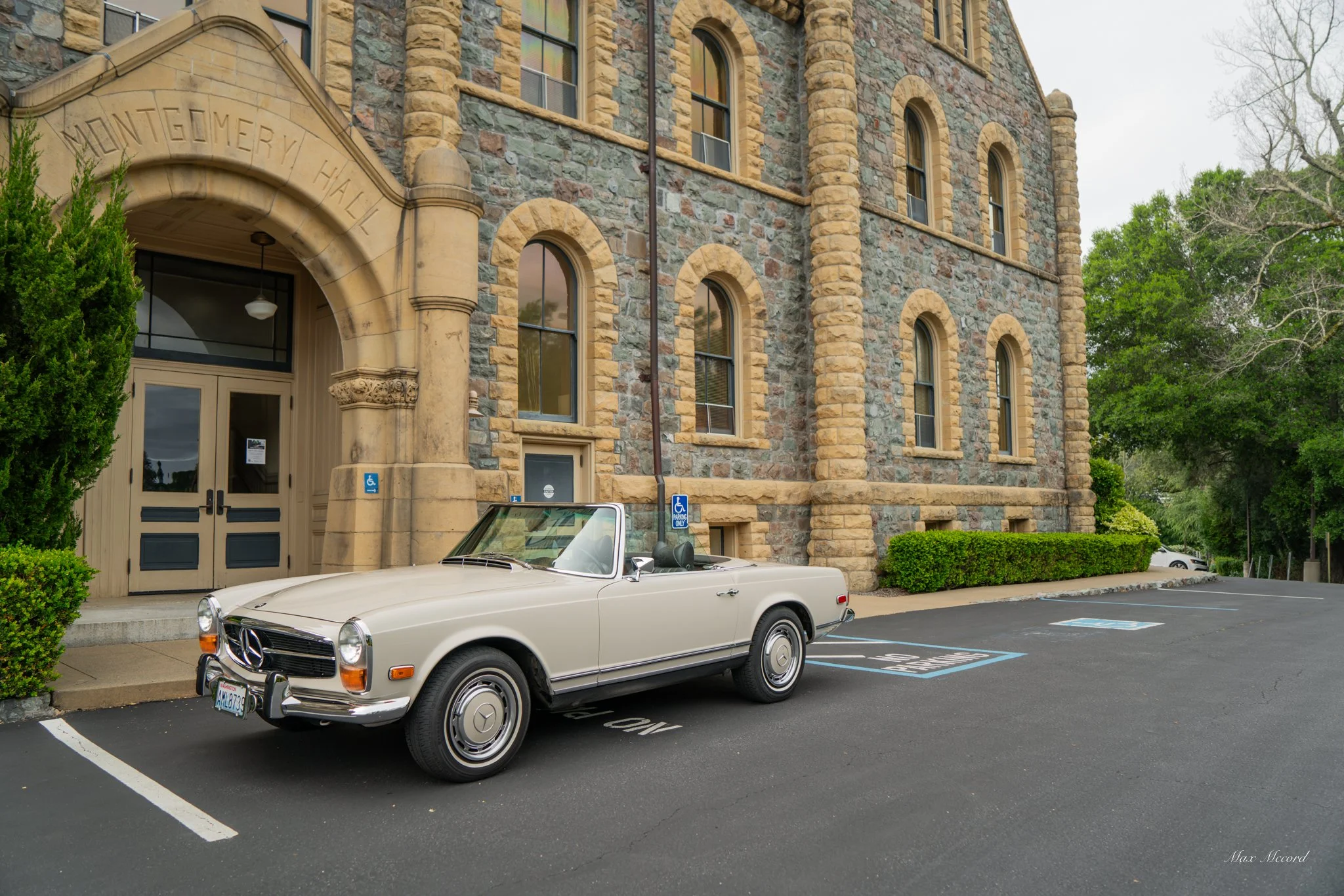 A vintage beige convertible Mercedes-Benz car parked in front of Montgomery Hall, a stone building with arched windows and an arched entrance, surrounded by greenery and a few other parked cars.