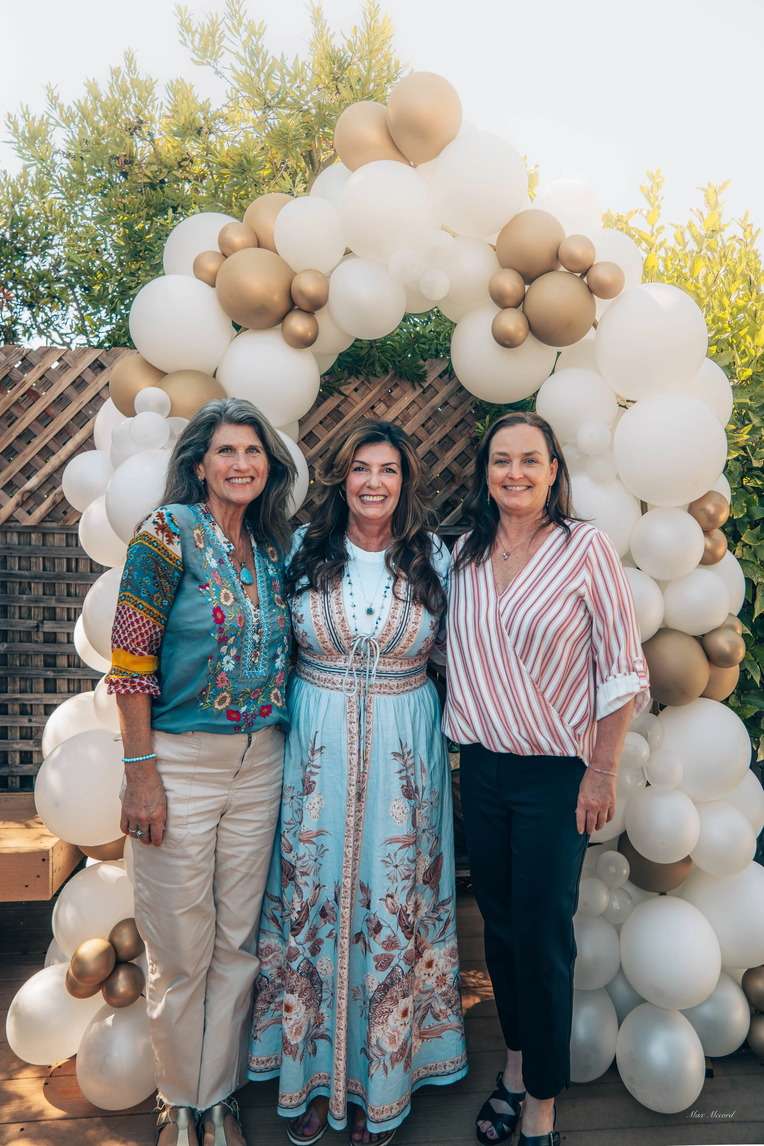 Three women standing side by side in front of a balloon arch at an outdoor event, smiling at the camera.