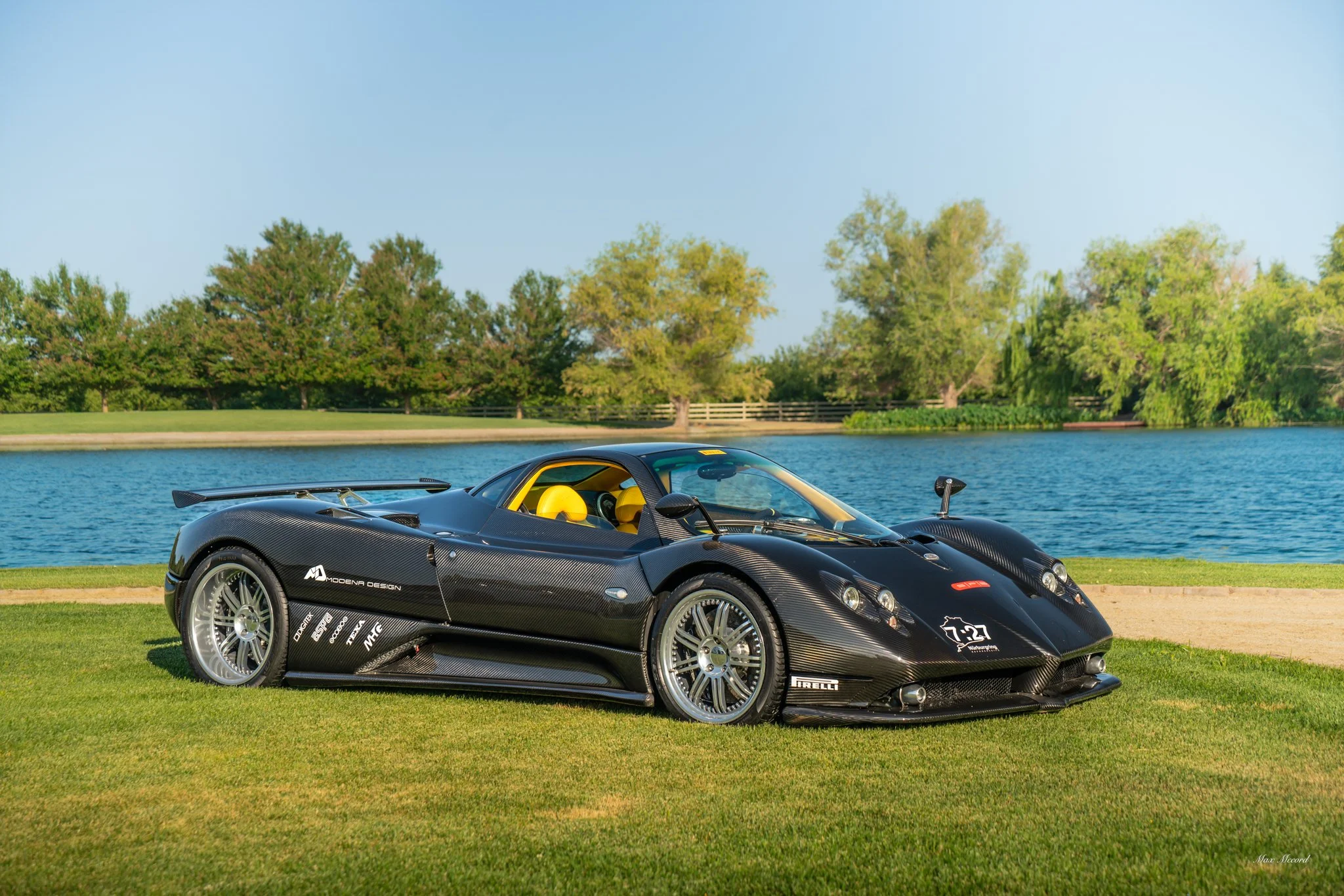 A black race car with yellow interior parked on grass near a lake with trees in the background.