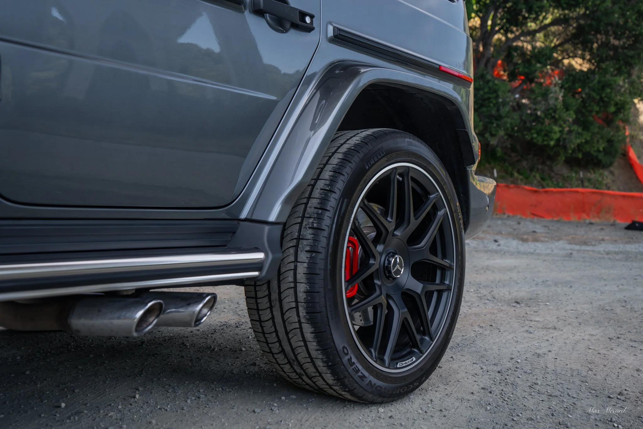 Close-up of a grey Mercedes-Benz G-Class SUV's rear wheel with black rims, mounted with Pirelli Scorpion Zero tires, showing red brake calipers, parked on gravel with a background of trees and orange construction barriers.