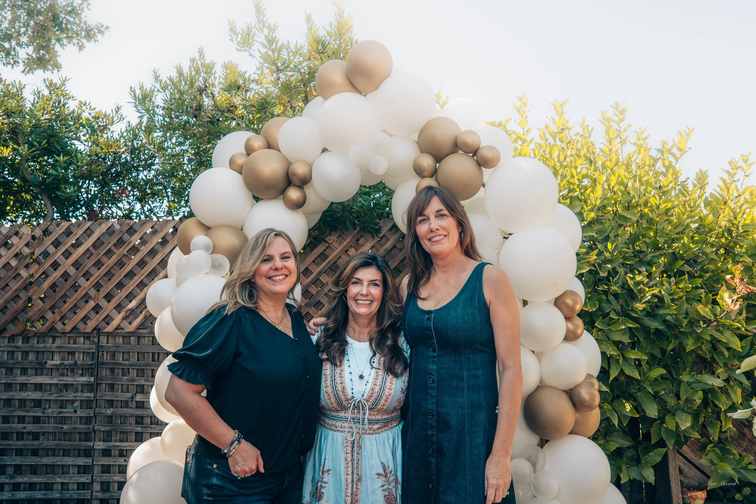 Three women standing together outdoors near a balloon arch with white and gold balloons, smiling at the camera.