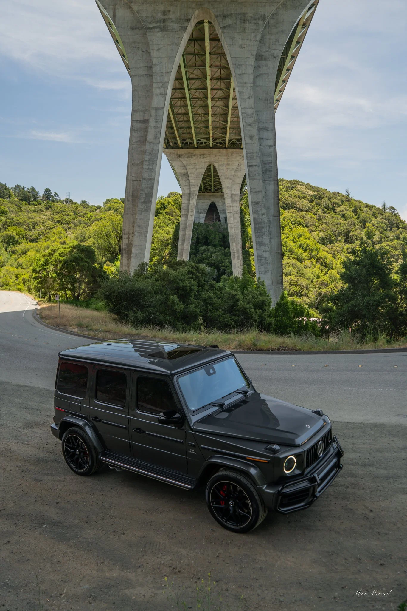 A black Mercedes-Benz G-Class SUV parked on a dirt road beneath a large concrete bridge surrounded by green trees and hills.