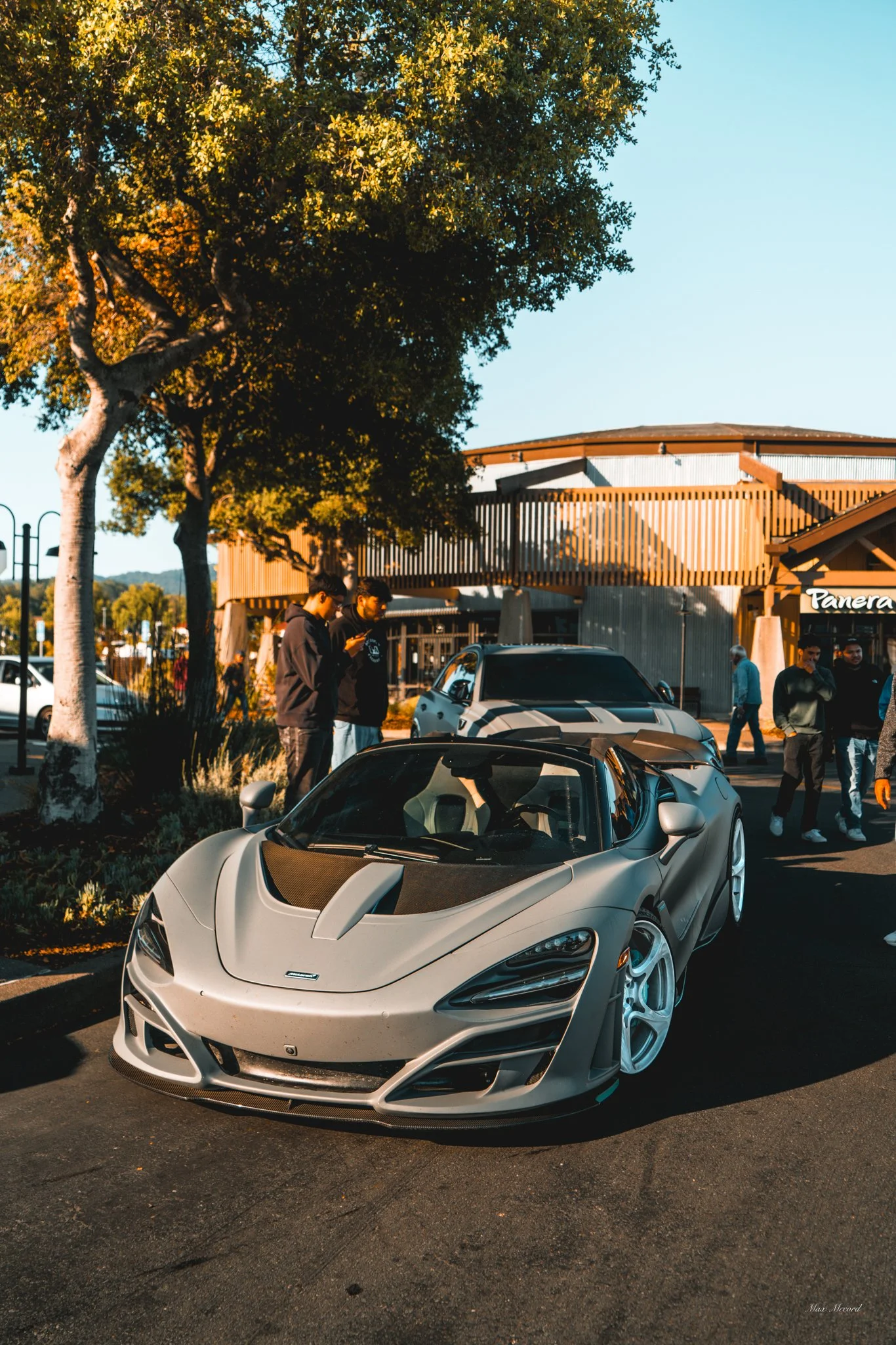 A silver sports car parked in a parking lot with people around, trees, and a building in the background during the daytime.