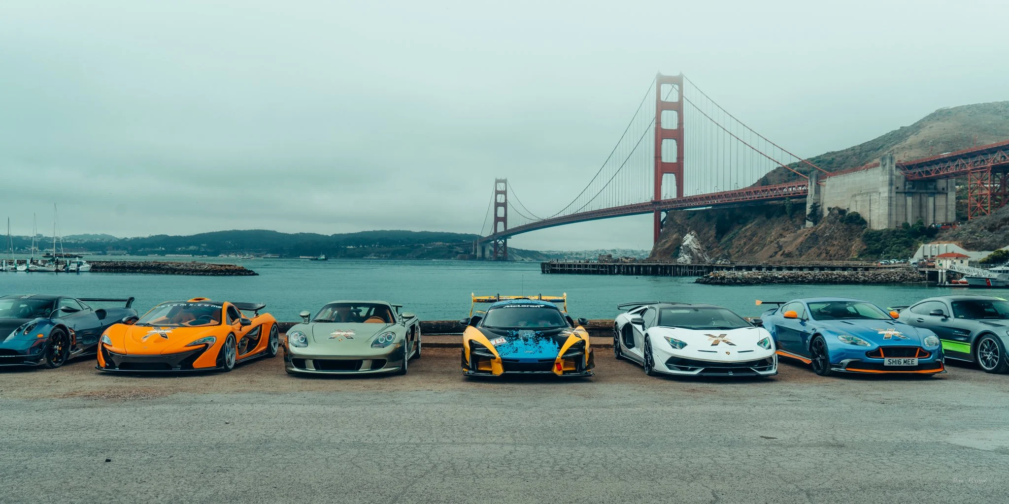 Five high-performance sports cars parked in front of the Golden Gate Bridge in San Francisco, with water and hills in the background.