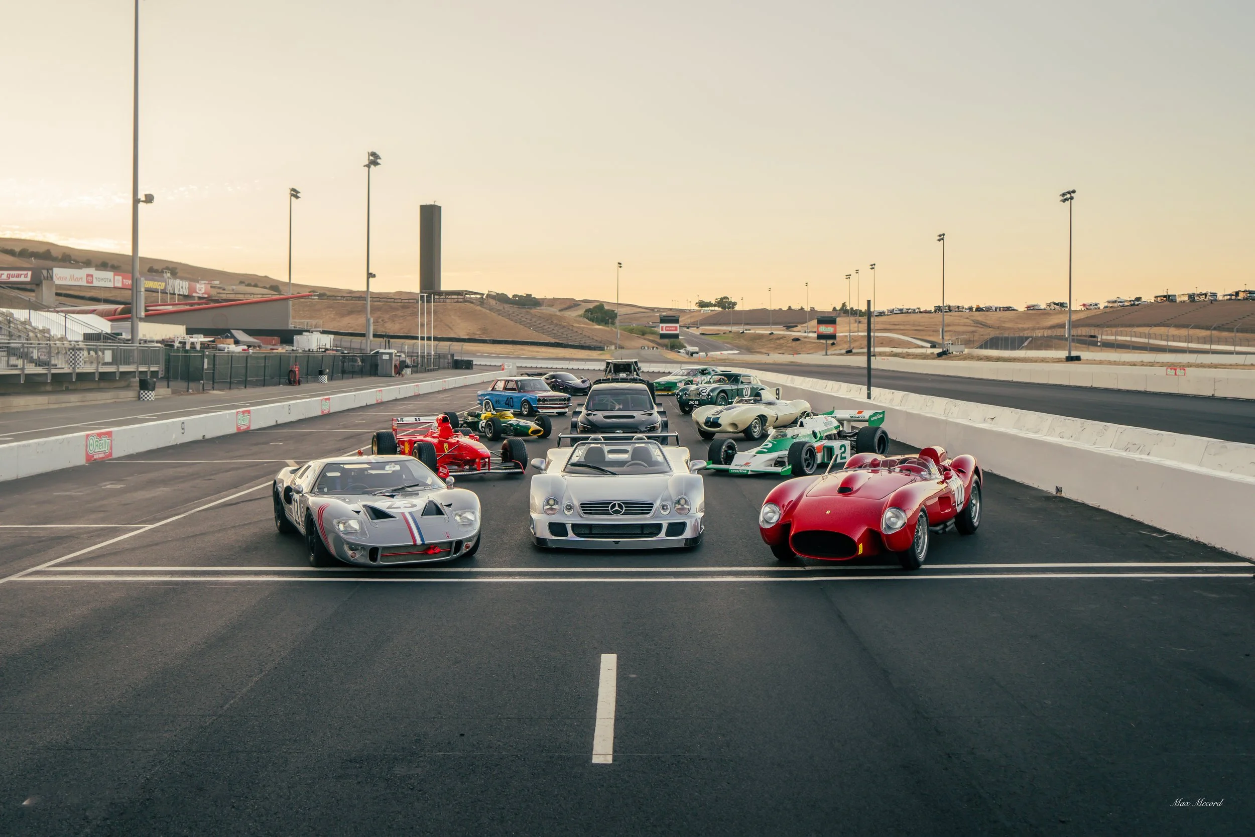 A lineup of classic racing cars on a racetrack, with a variety of vintage models including Mercedes, Ferrari, and other sports cars, set against a backdrop of empty stands and a hilly landscape during sunset.