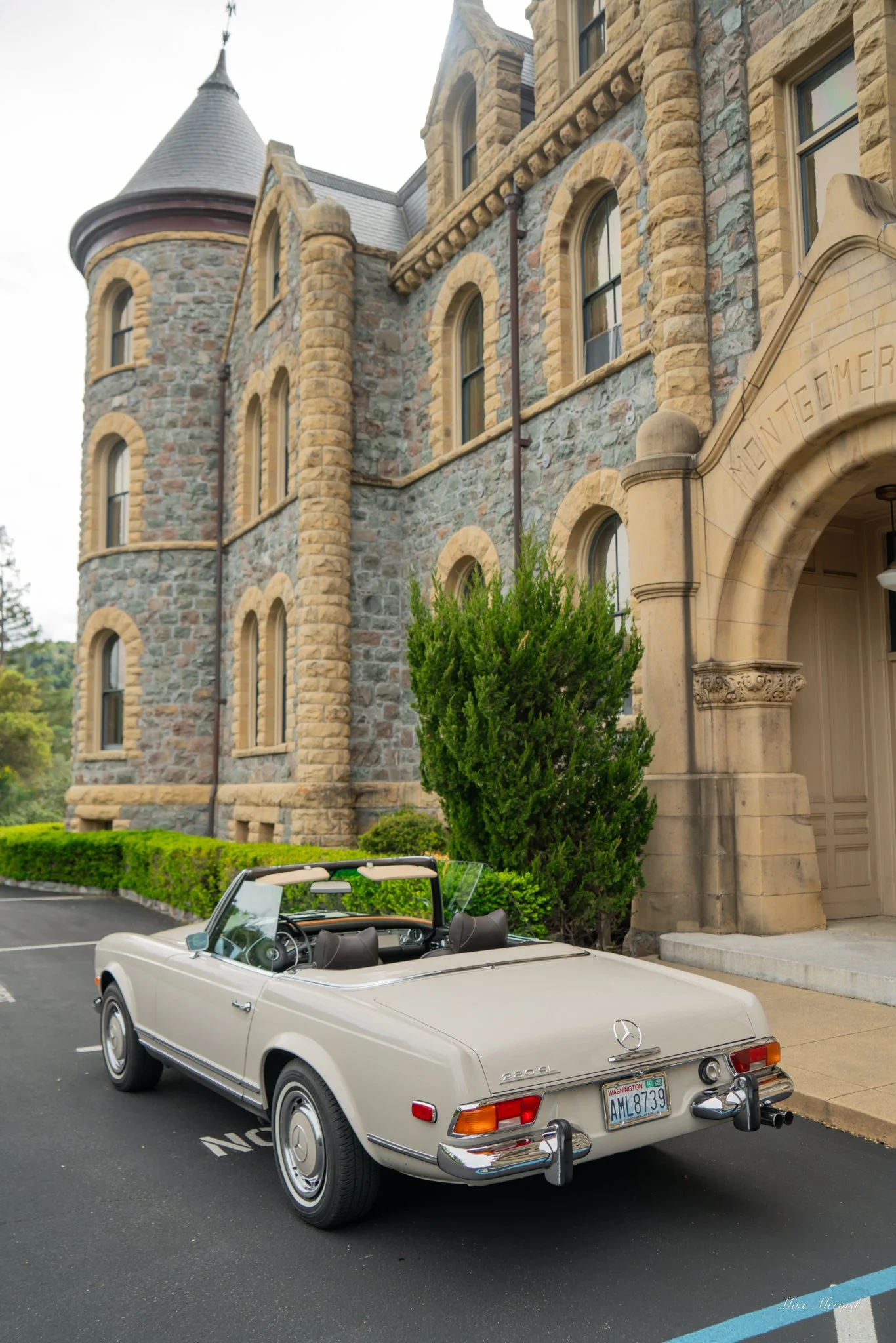 A vintage white Mercedes-Benz 280SL convertible parked in front of a stone castle-like building with an arched entrance and multiple windows.