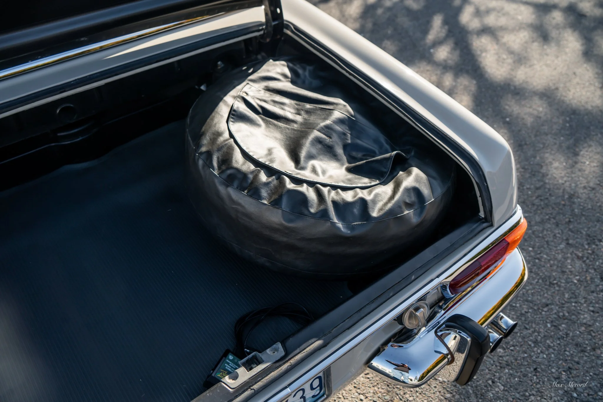 The trunk of a vintage silver car with a spare tire in a black cover, parked on a paved surface.