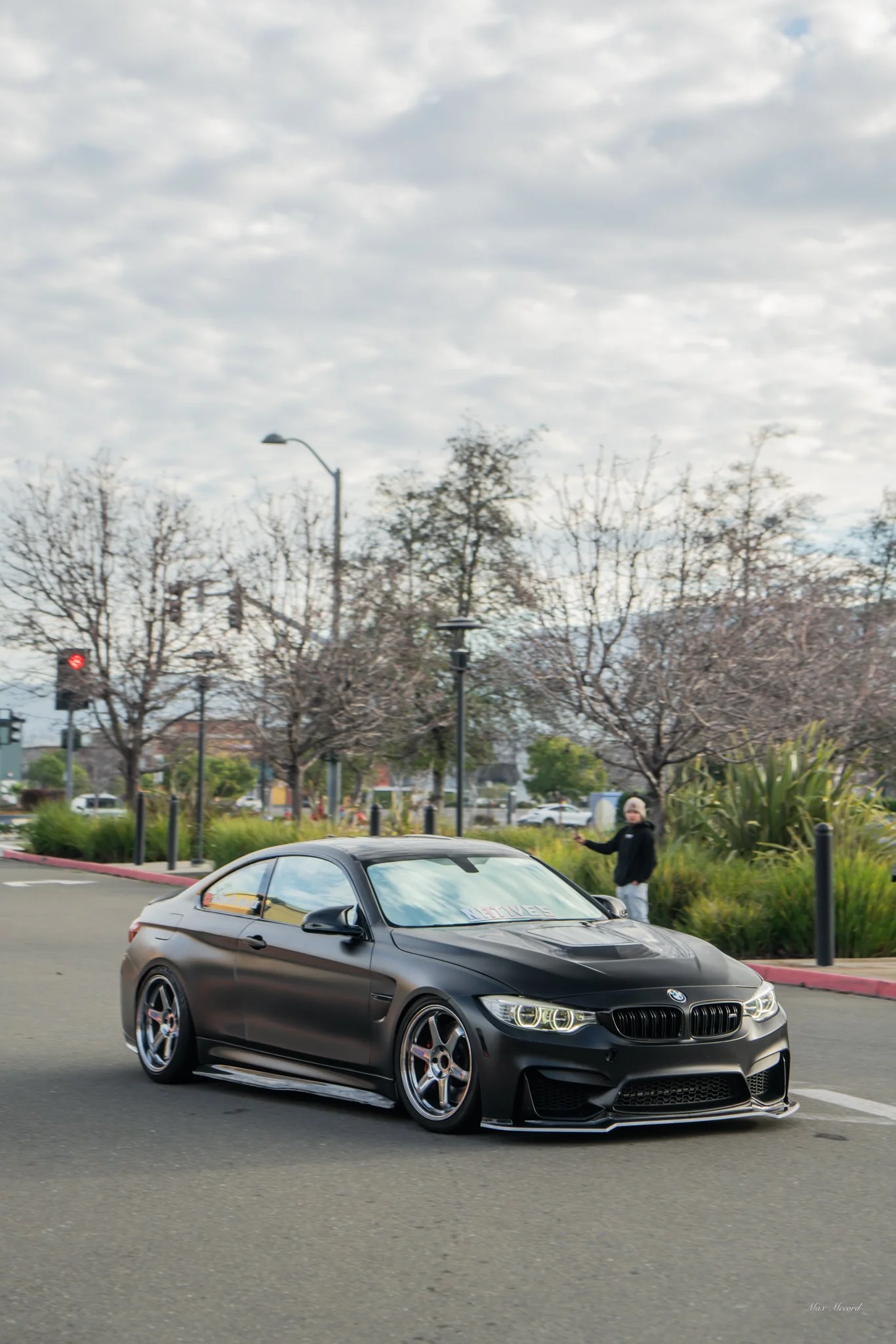 A black BMW sports car driving on a city street with a person standing on the sidewalk and trees in the background.