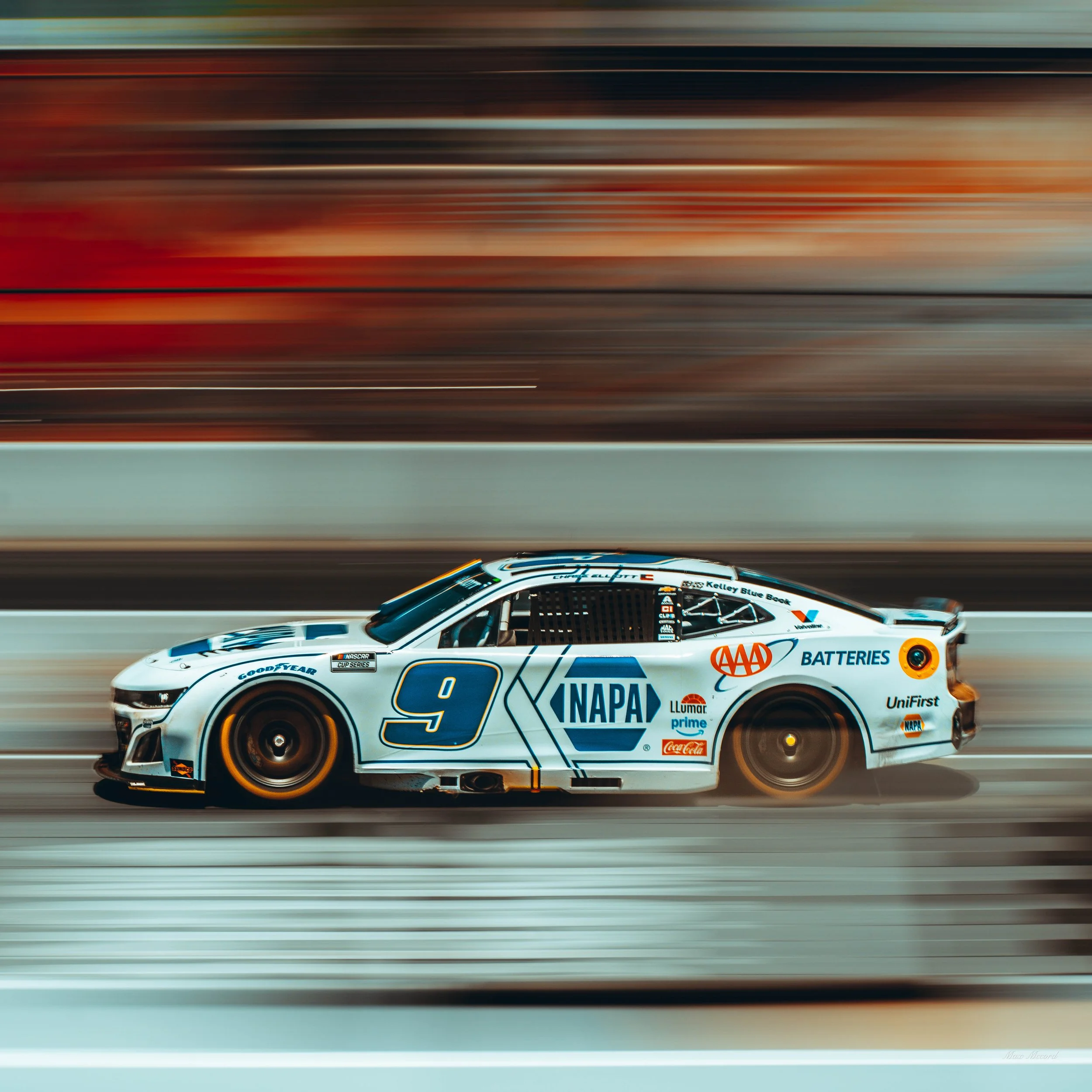 A NASCAR race car with the number 9, sponsored by NAPA, racing on a track at high speed during sunset, with motion blur in the background.
