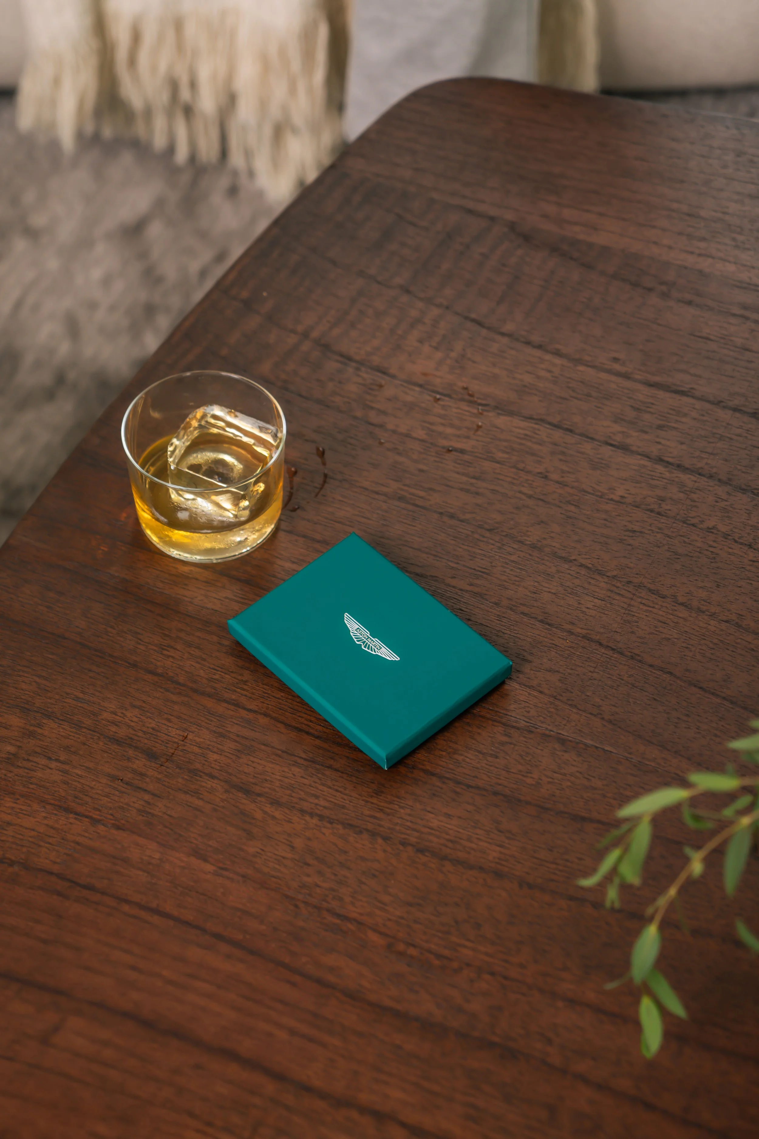 A wooden table with a glass of whiskey and a teal box with a white logo on it.