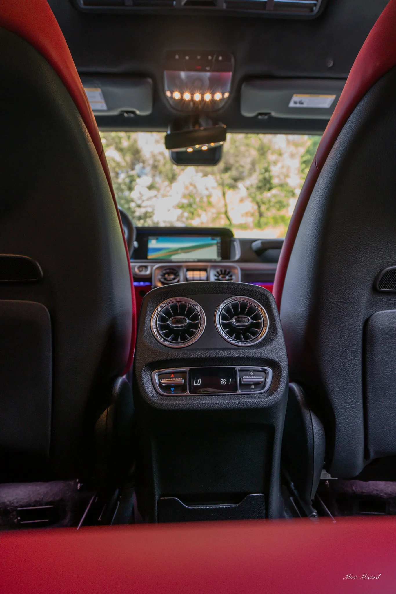 Interior of a luxury car viewed from the back seat, showing the air conditioning vents and controls between the driver and passenger seats, with a digital navigation screen visible through the windshield, and trees visible outside.