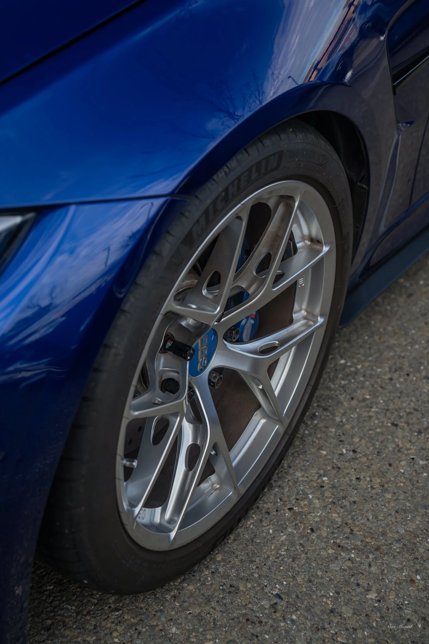 Close-up of a blue sports car's front wheel and part of the fender, with a modern alloy rim and tire.