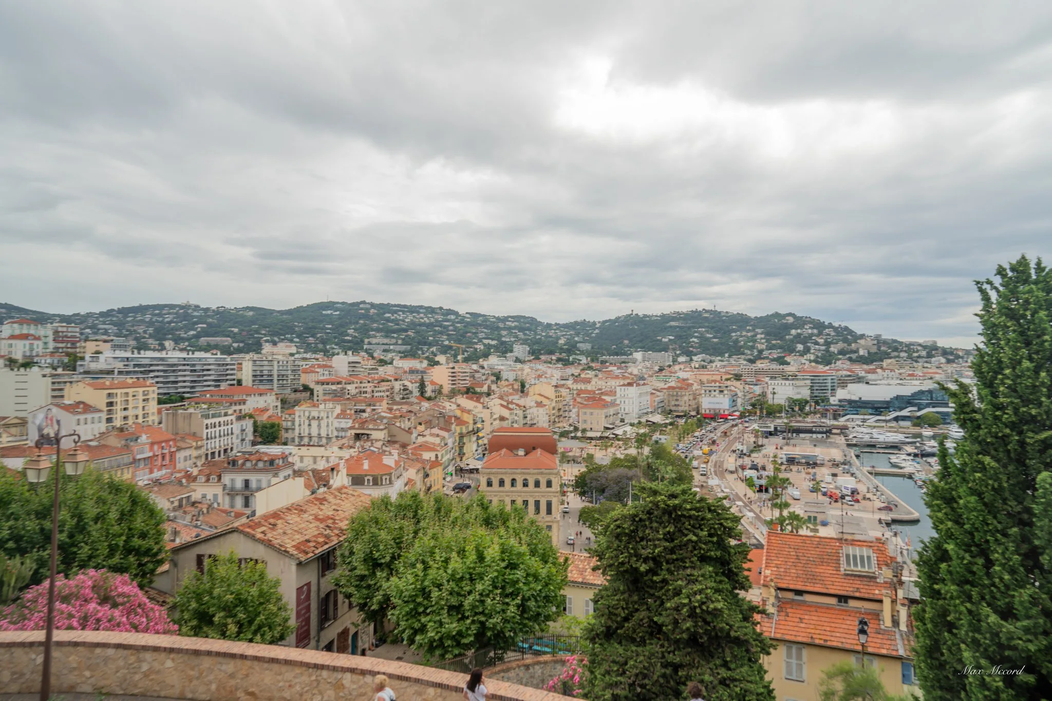 View of a coastal city with a harbor, numerous buildings with red-tiled roofs, some taller modern structures, hills in the background, and cloudy skies overhead.