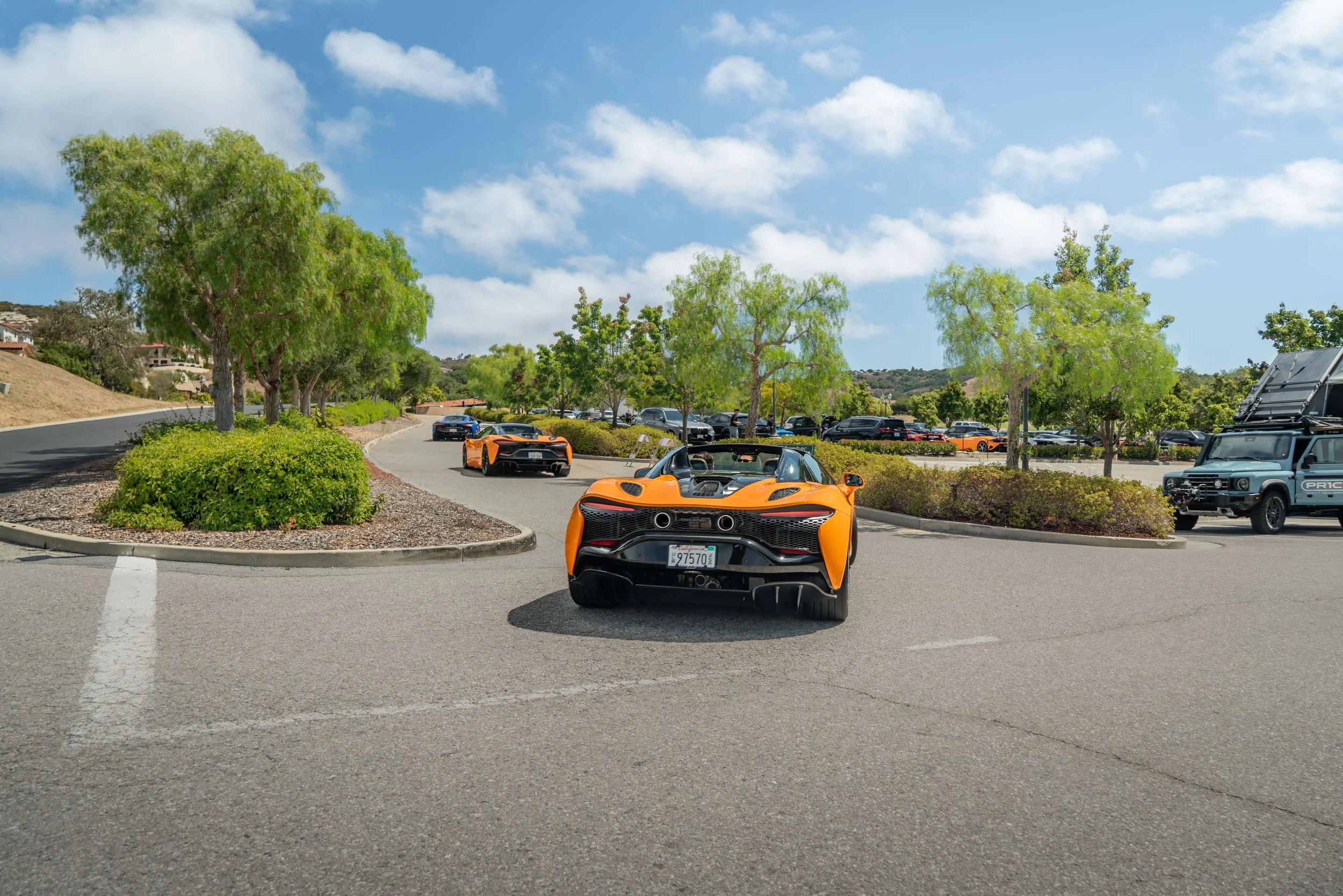 Orange sports cars parked along a winding parking lot lined with trees, under a partly cloudy sky.
