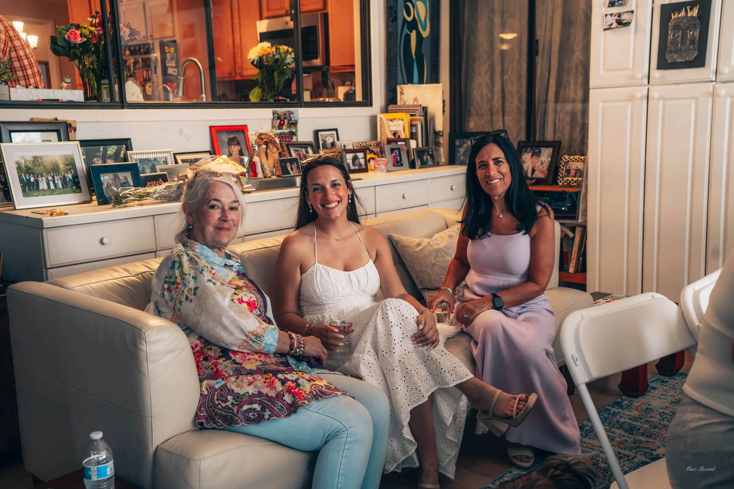 Three women sitting on a cream-colored couch in a living room, smiling at the camera. The woman on the left has gray hair and is wearing a colorful, floral blouse. The woman in the middle has long dark hair and is dressed in a white eyelet dress, hol