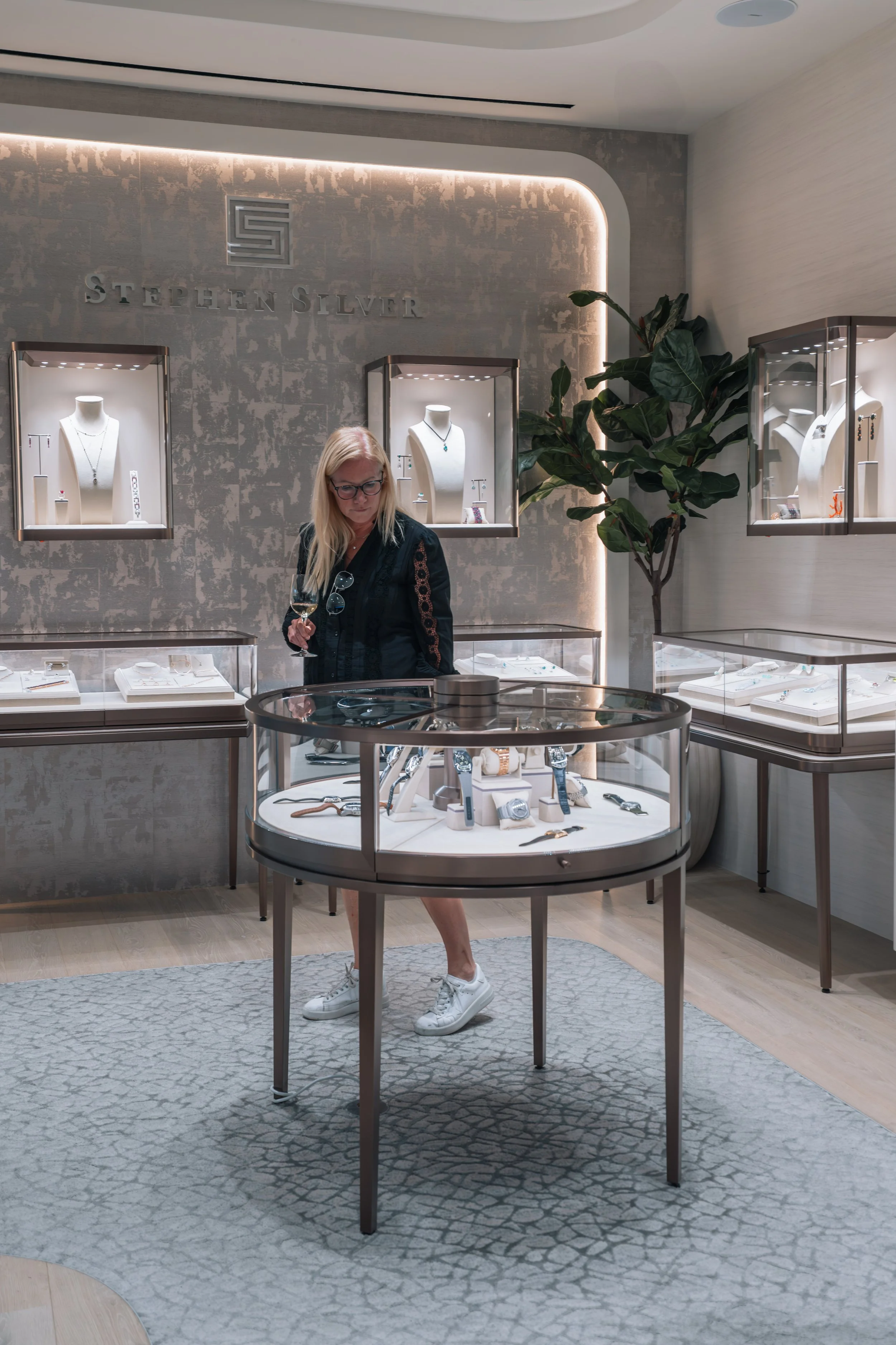 Woman shopping for jewelry in a store, holding a glass of wine, with jewelry displays behind her and a potted plant nearby.