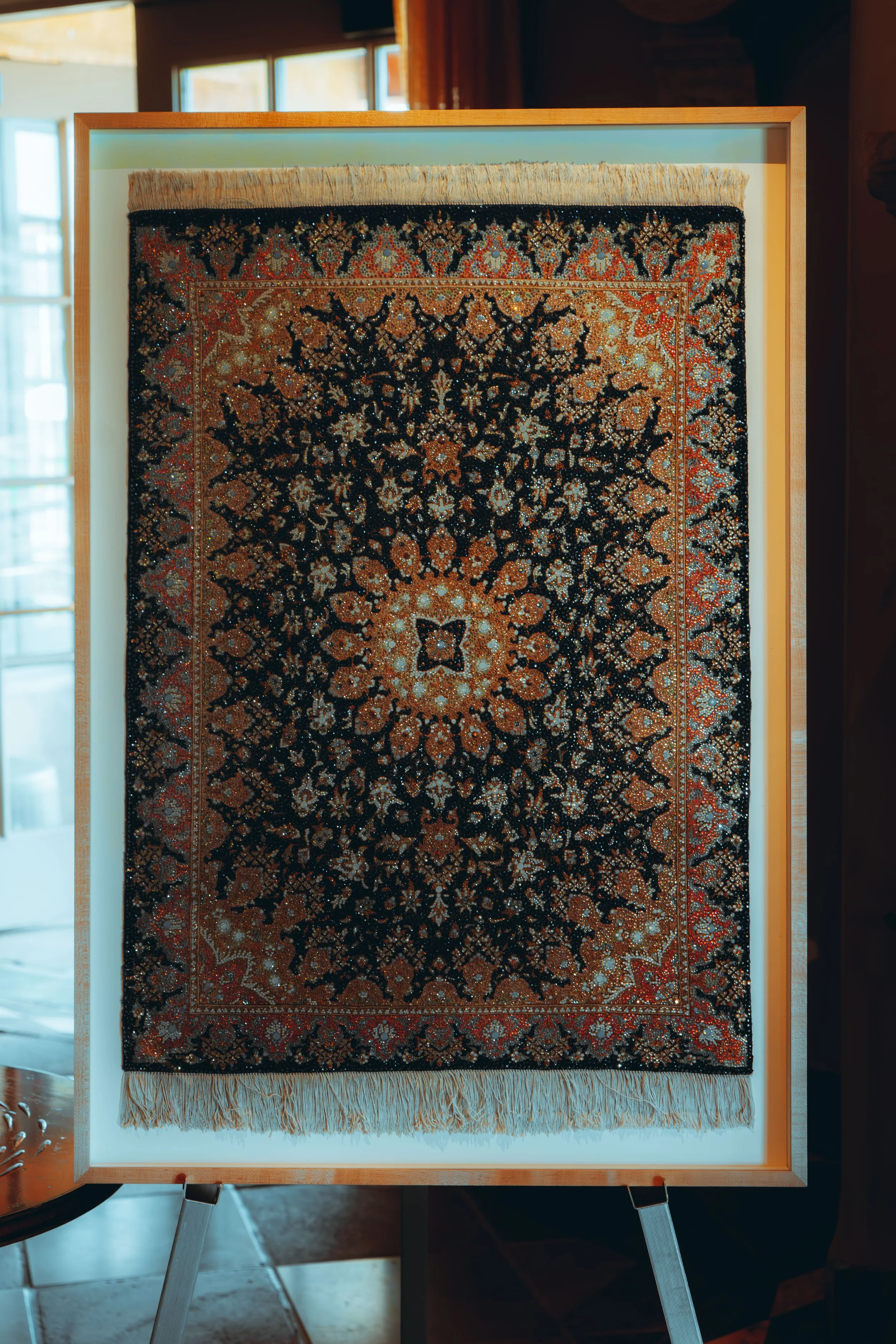 A framed Persian rug with intricate floral and geometric patterns in black, orange, beige, and blue, displayed upright on a lighted stand indoors.