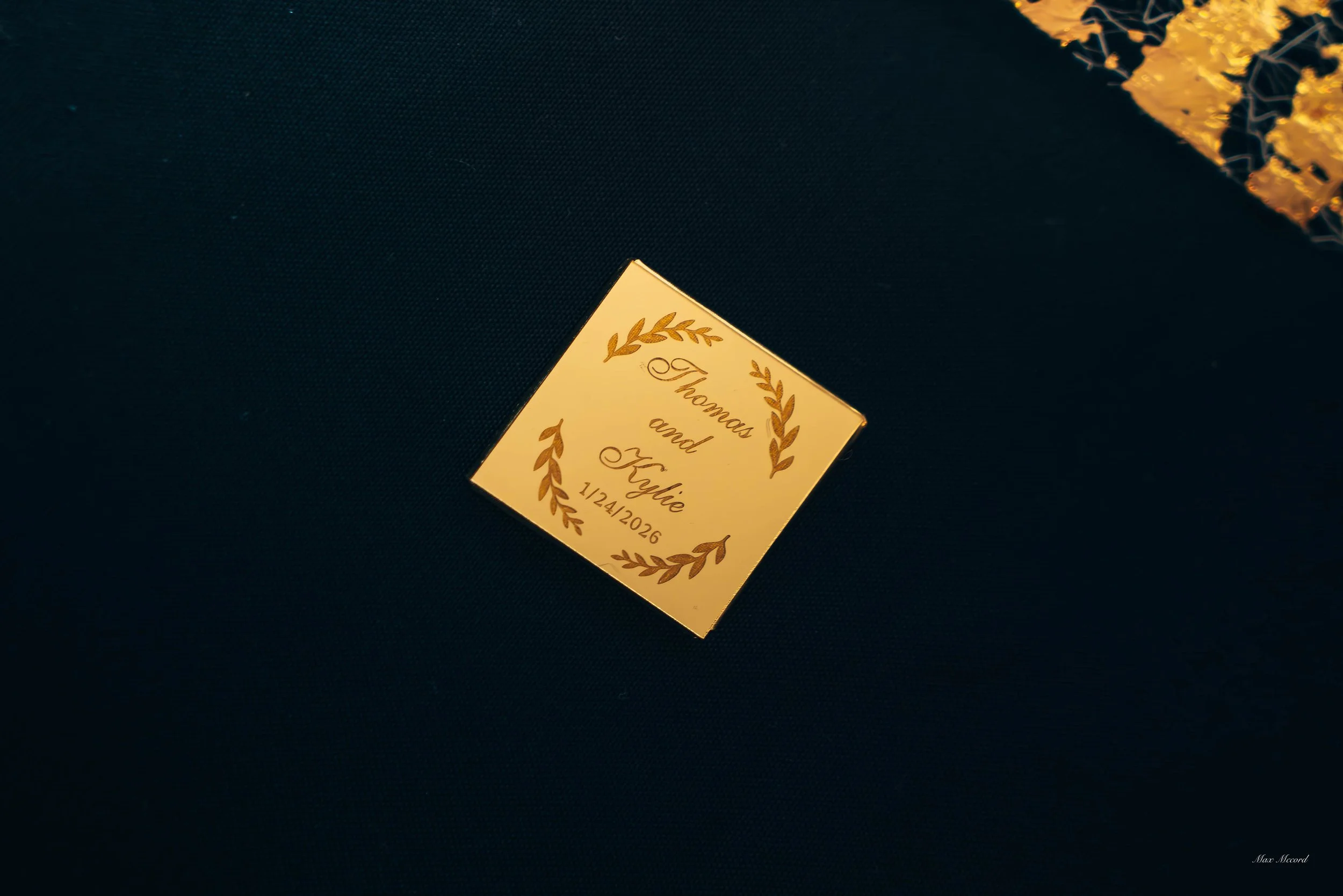 Wedding table card with gold lettering that says 'Thomas and Kylie, 1/24/2026' surrounded by a gold leaf border, on a dark table.