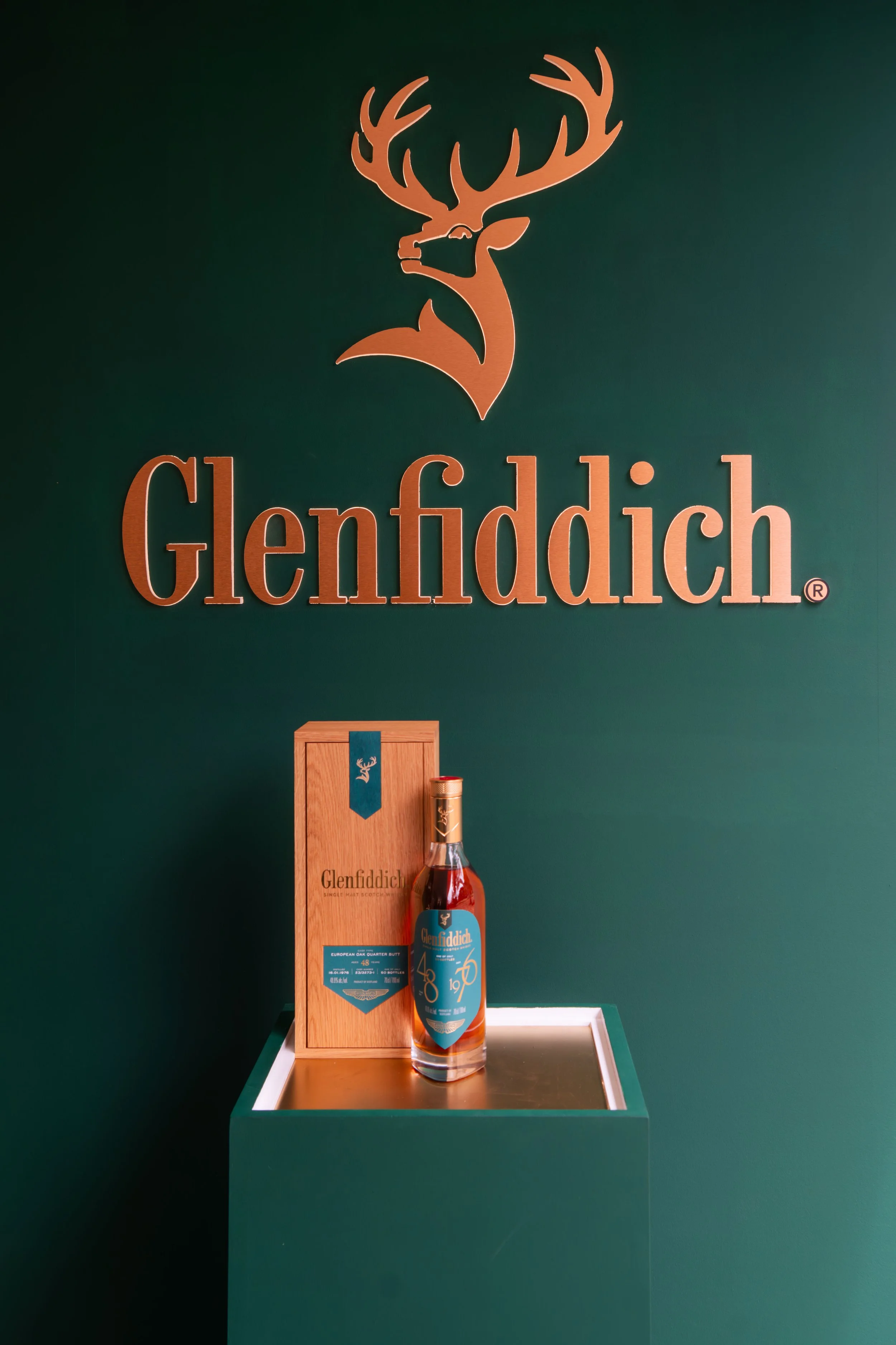 A bottle of Glenfiddich whisky displayed on a stand with a wooden box, set against a green wall with the Glenfiddich logo and brand name.