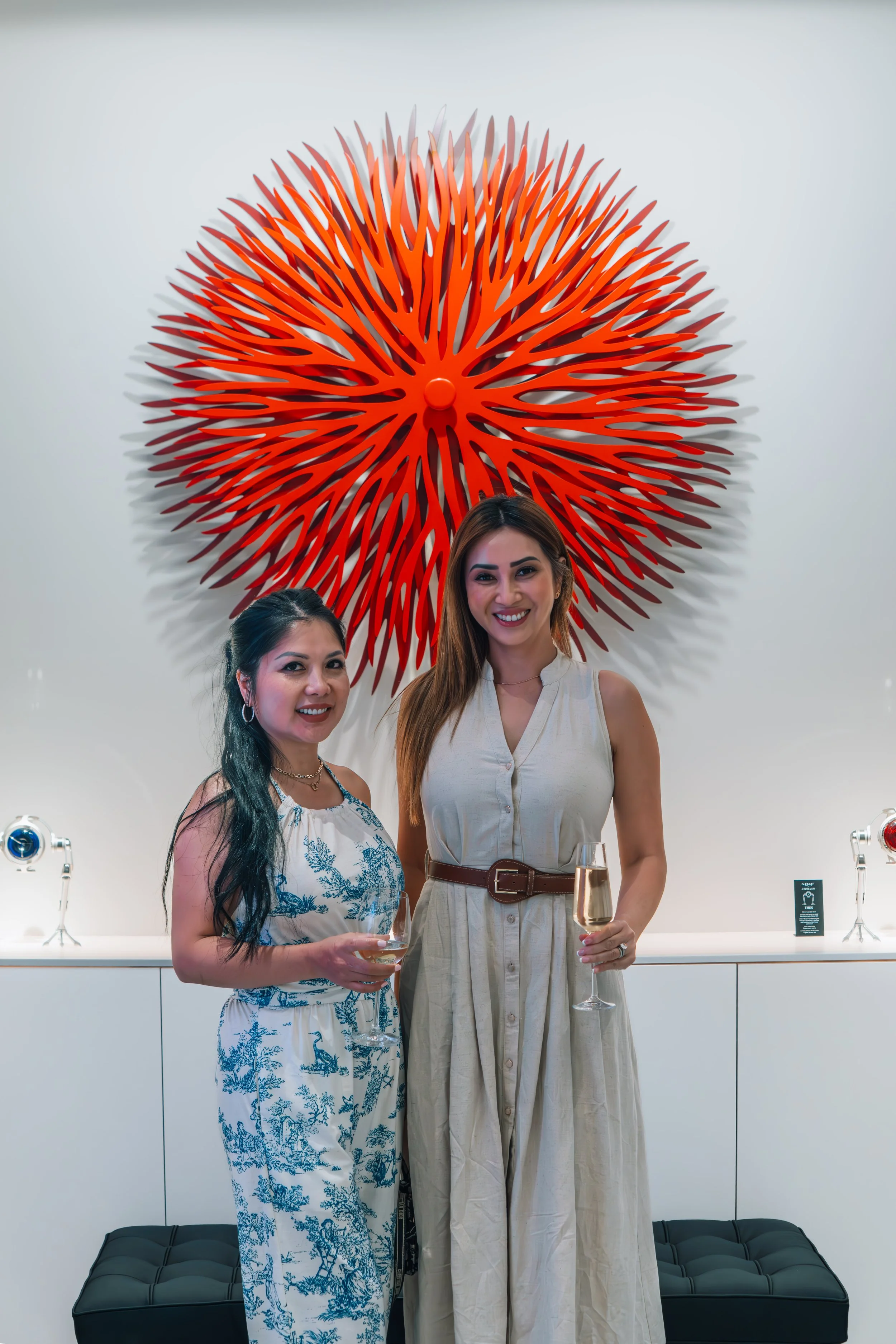 Two women standing in front of a large orange art piece on a white wall, holding glasses of champagne and smiling.