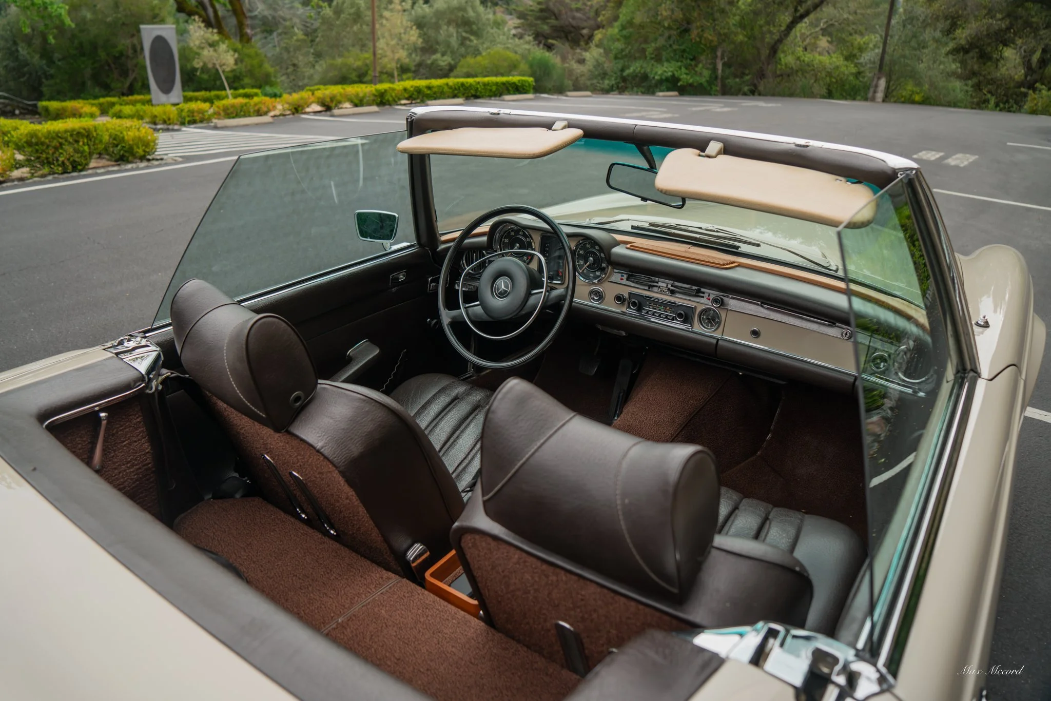 Interior of a vintage convertible car with brown leather seats and a classic dashboard, parked in an outdoor lot with greenery in the background.