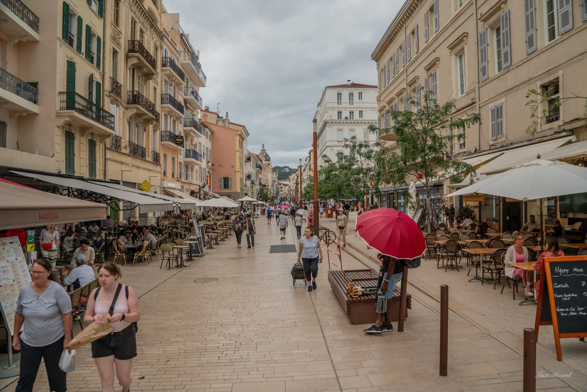 A bustling city street with outdoor cafes, people walking, and buildings with shuttered windows under an overcast sky.