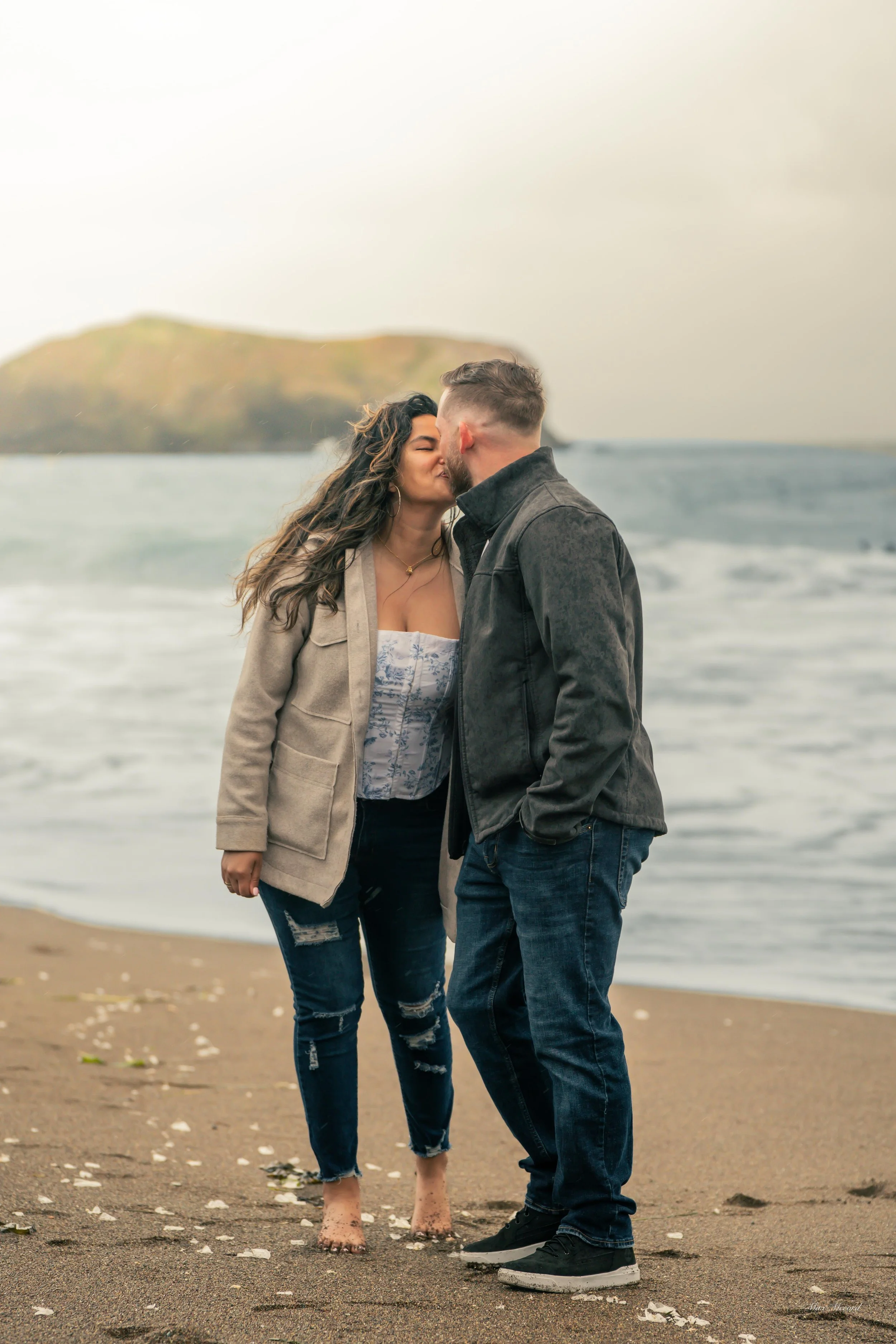 A romantic couple sharing a kiss on the beach with ocean waves and a hill in the background.