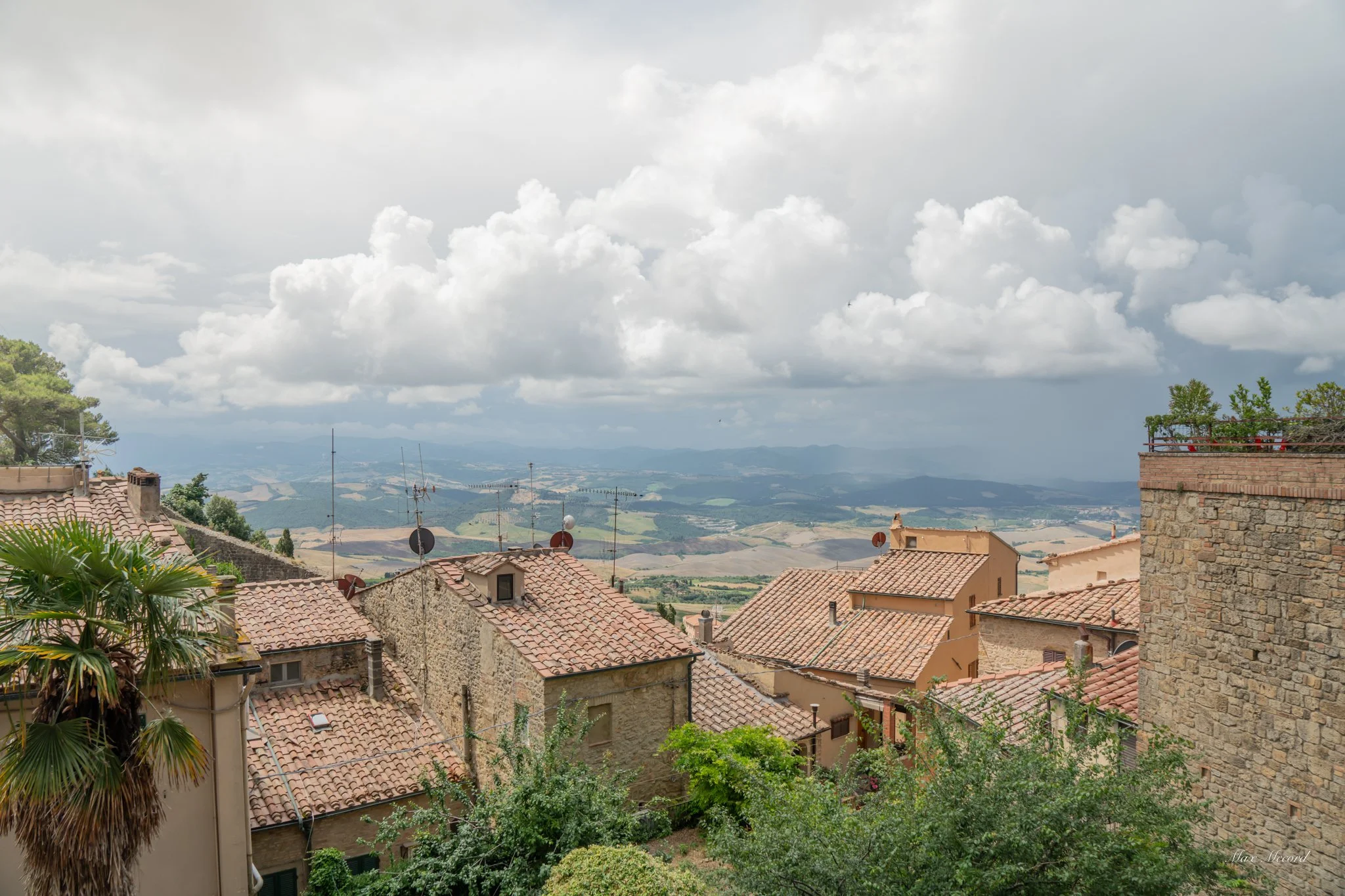 View of rooftops with terracotta tiles and antennas in a hillside town, with green trees and rolling hills under a cloudy sky in the background.