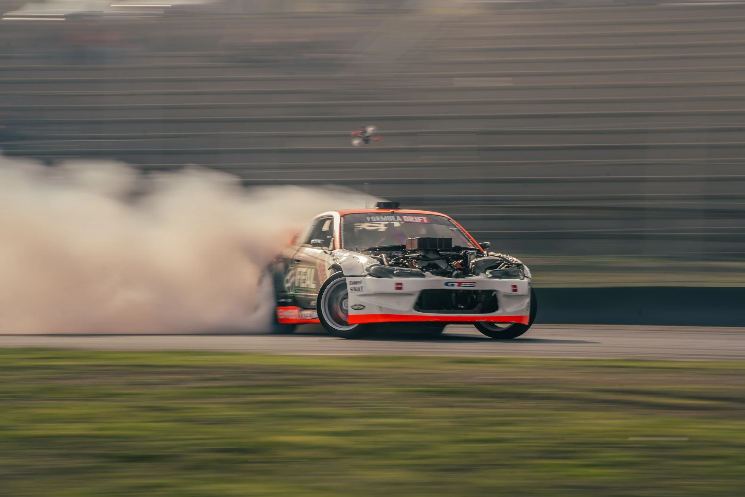 A white racing car with red and black accents drifting on a racetrack, creating tire smoke. The car has an exposed engine and signage indicating it's participating in a drift event.