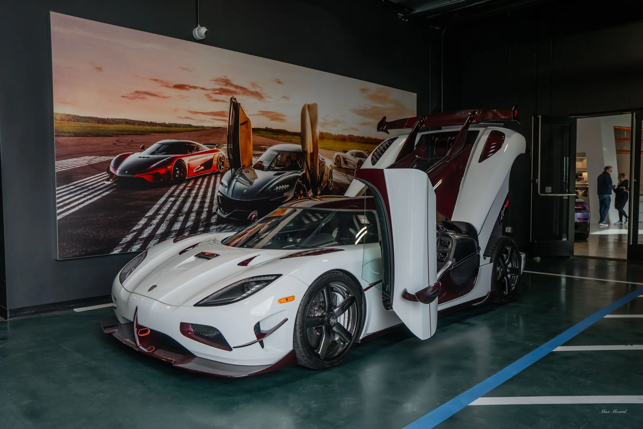 A white and burgundy sports car with its doors and roof open inside a showroom, with a large photo of racing cars on the wall behind it and two people in the background near a different car.