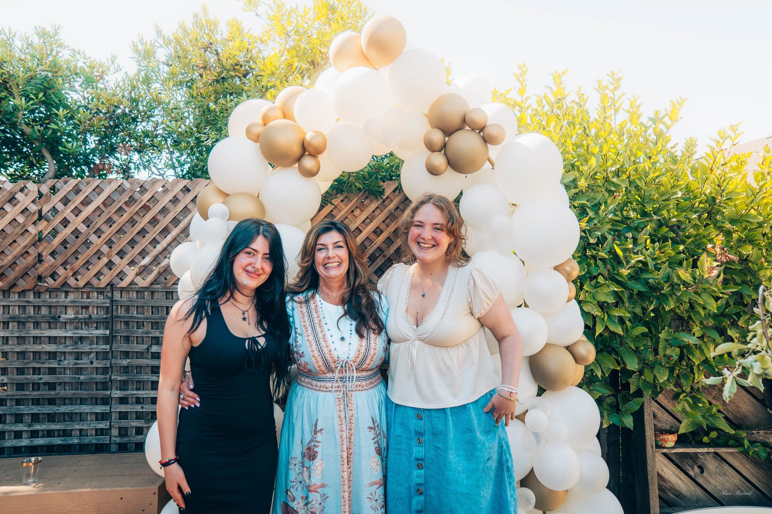 Three women standing together outdoors in front of a balloon arch decorated with white, beige, and gold balloons, with green trees and a wooden fence in the background, smiling and enjoying the occasion.