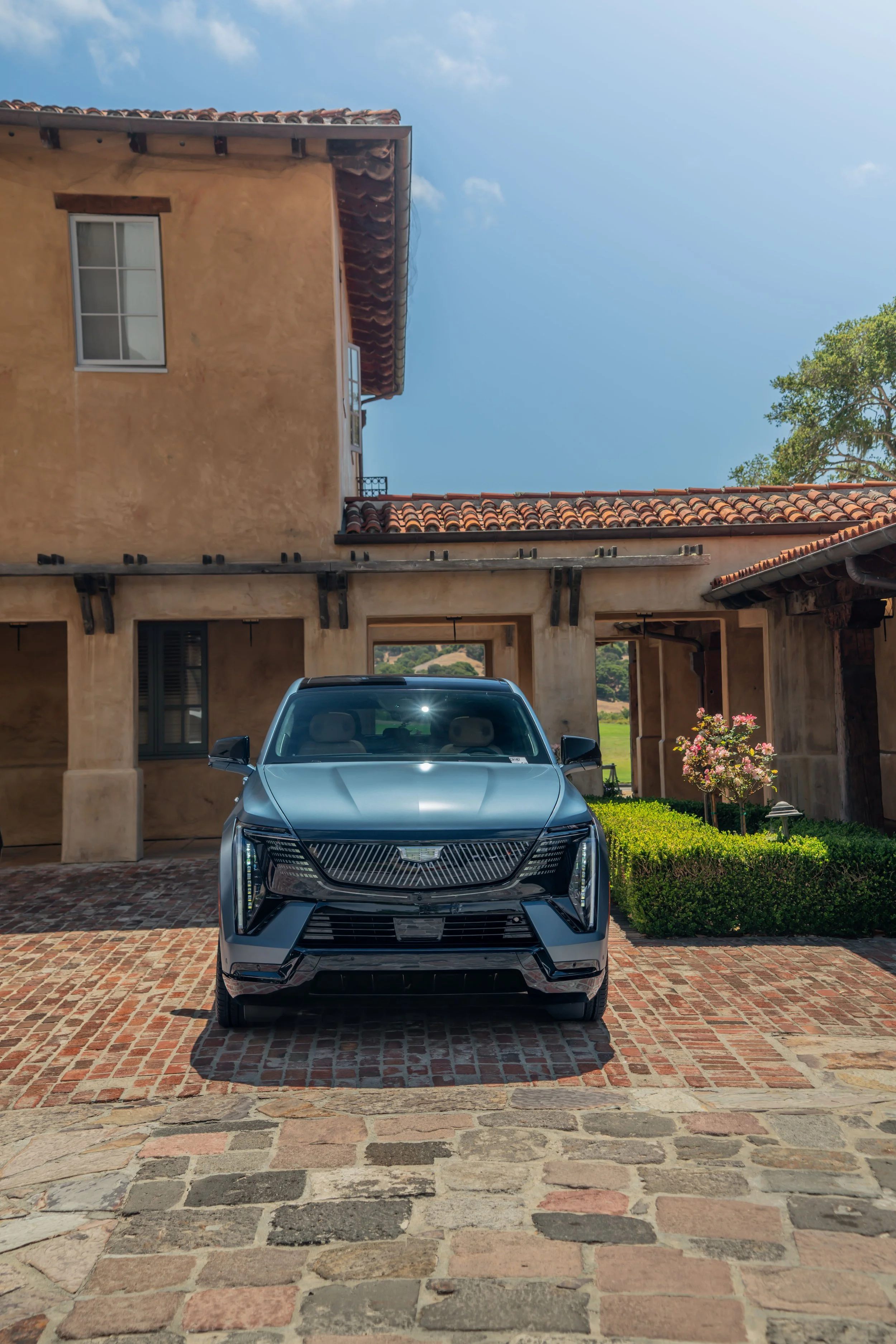 A black luxury SUV parked in front of a Mediterranean-style house with stucco walls and a red tile roof, under a blue sky with some clouds.