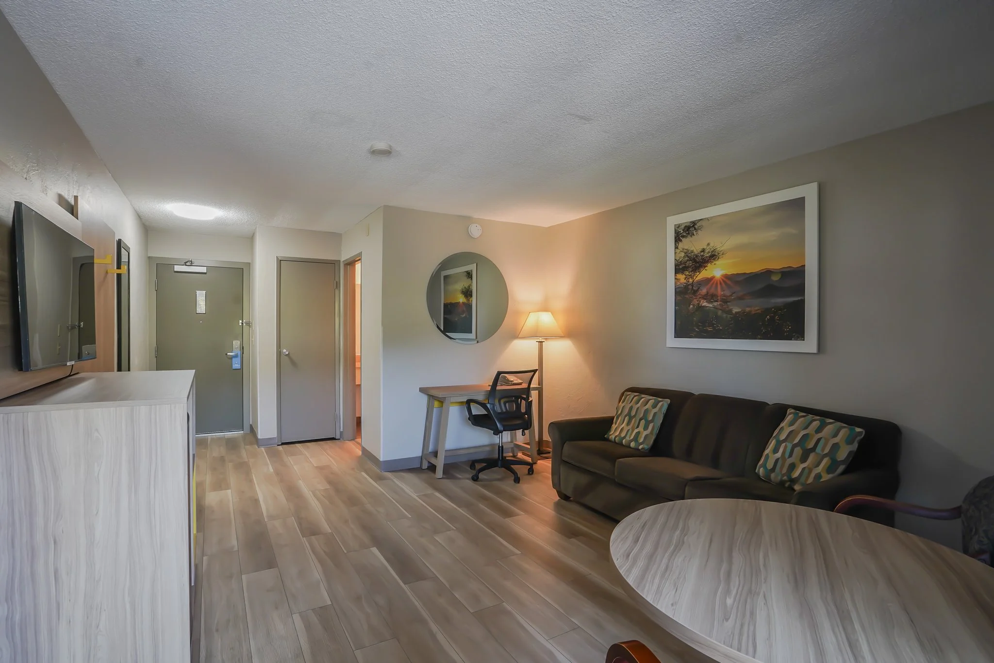 Living room with a TV on a stand, a green sofa with patterned pillows, a round wooden table, a small desk with a chair, a floor lamp, and framed landscape photo on wall.