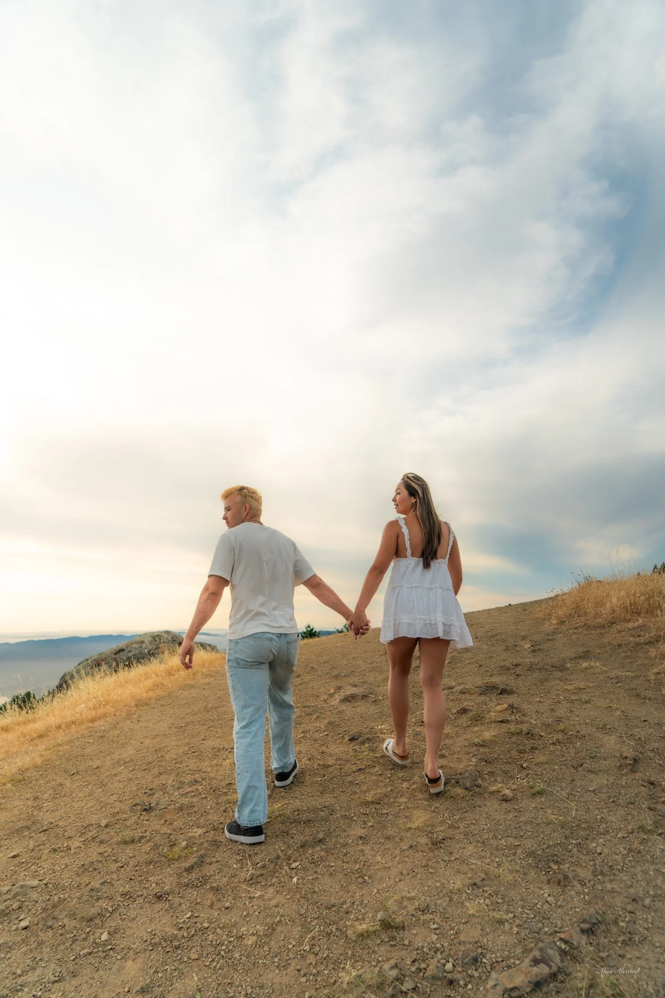 A young couple holding hands and walking up a dirt path on a hillside during sunset, with a cloudy sky overhead.
