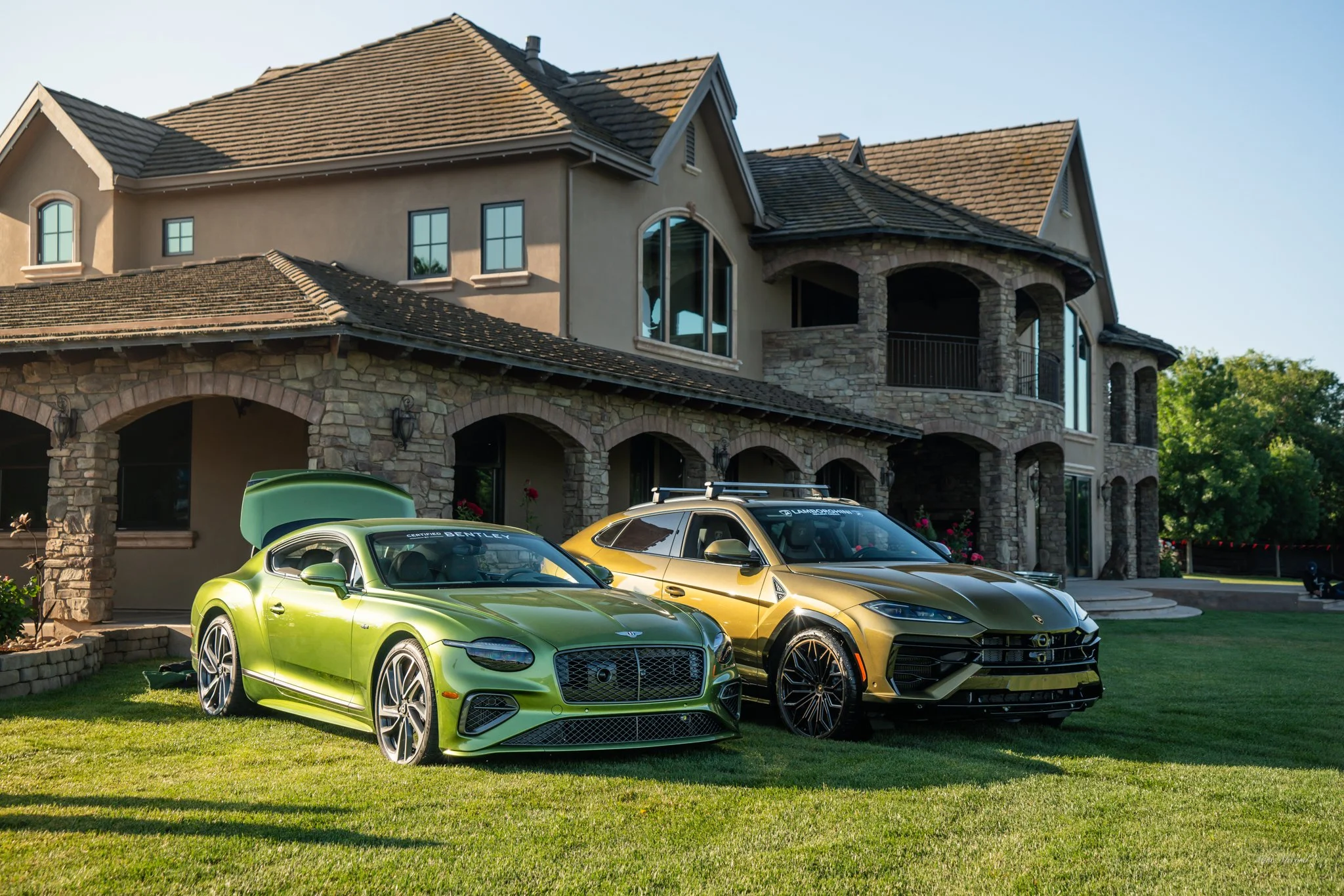 Two luxury cars, a green sedan and a gold SUV, parked on a lawn in front of a large stone and stucco house with multiple windows and a balcony, during daytime.