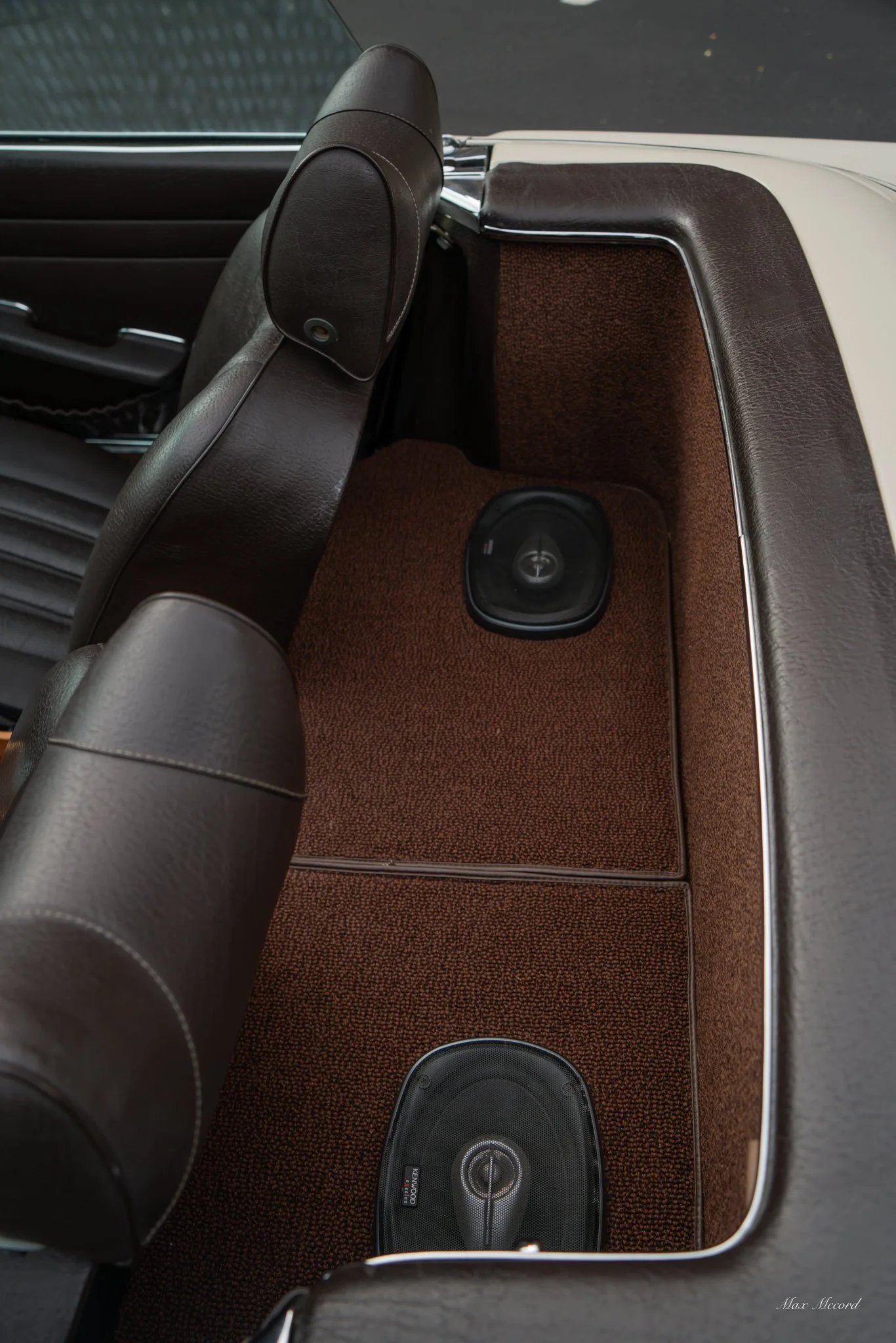 Interior of a vintage car showing the back of the front seats, a brown carpeted floor section with two oval speakers, and part of the passenger door with a handle and window.