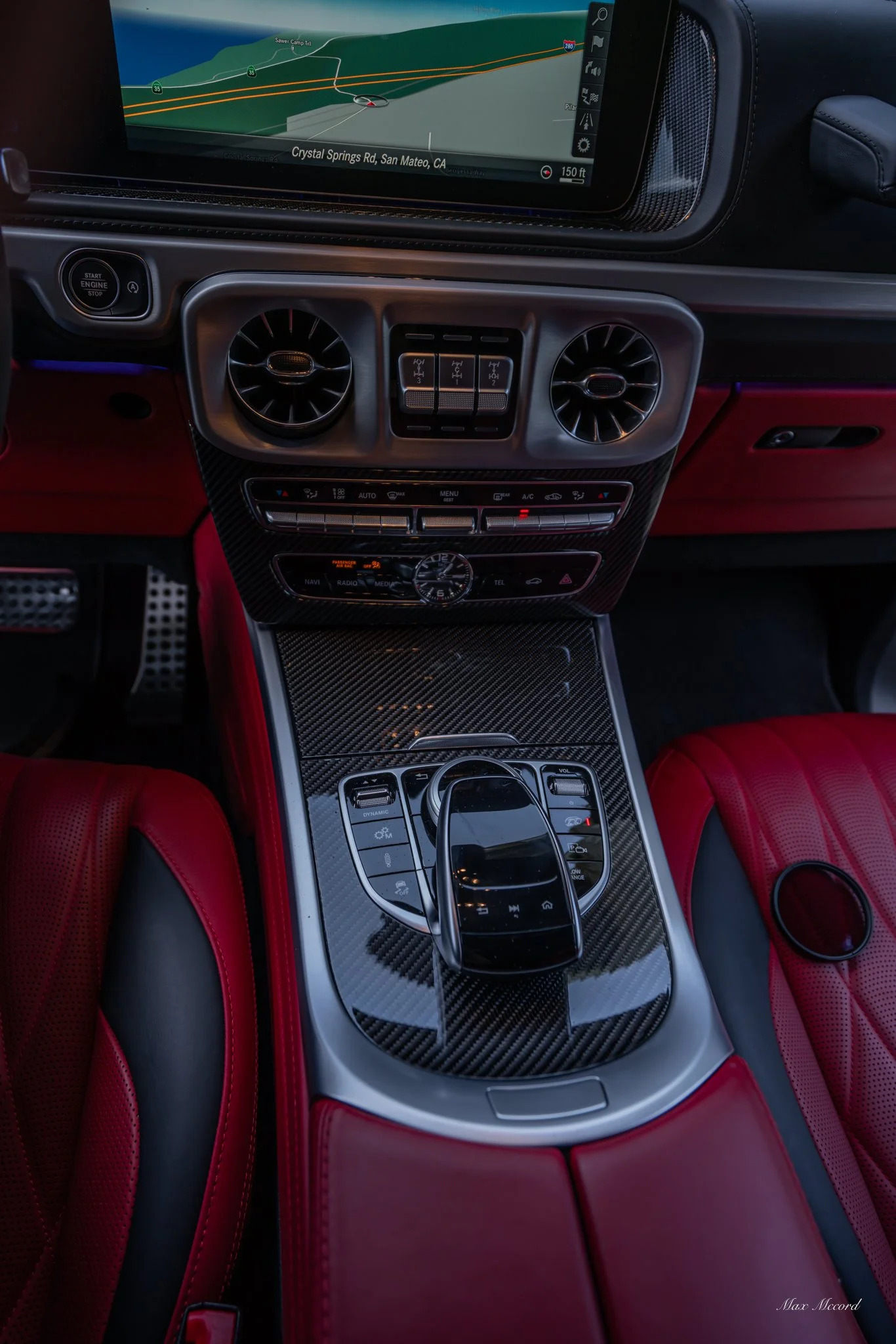 Interior of a luxury car dashboard showing a GPS navigation screen, climate controls, and a gear shift with a touchpad, all with red leather seats.