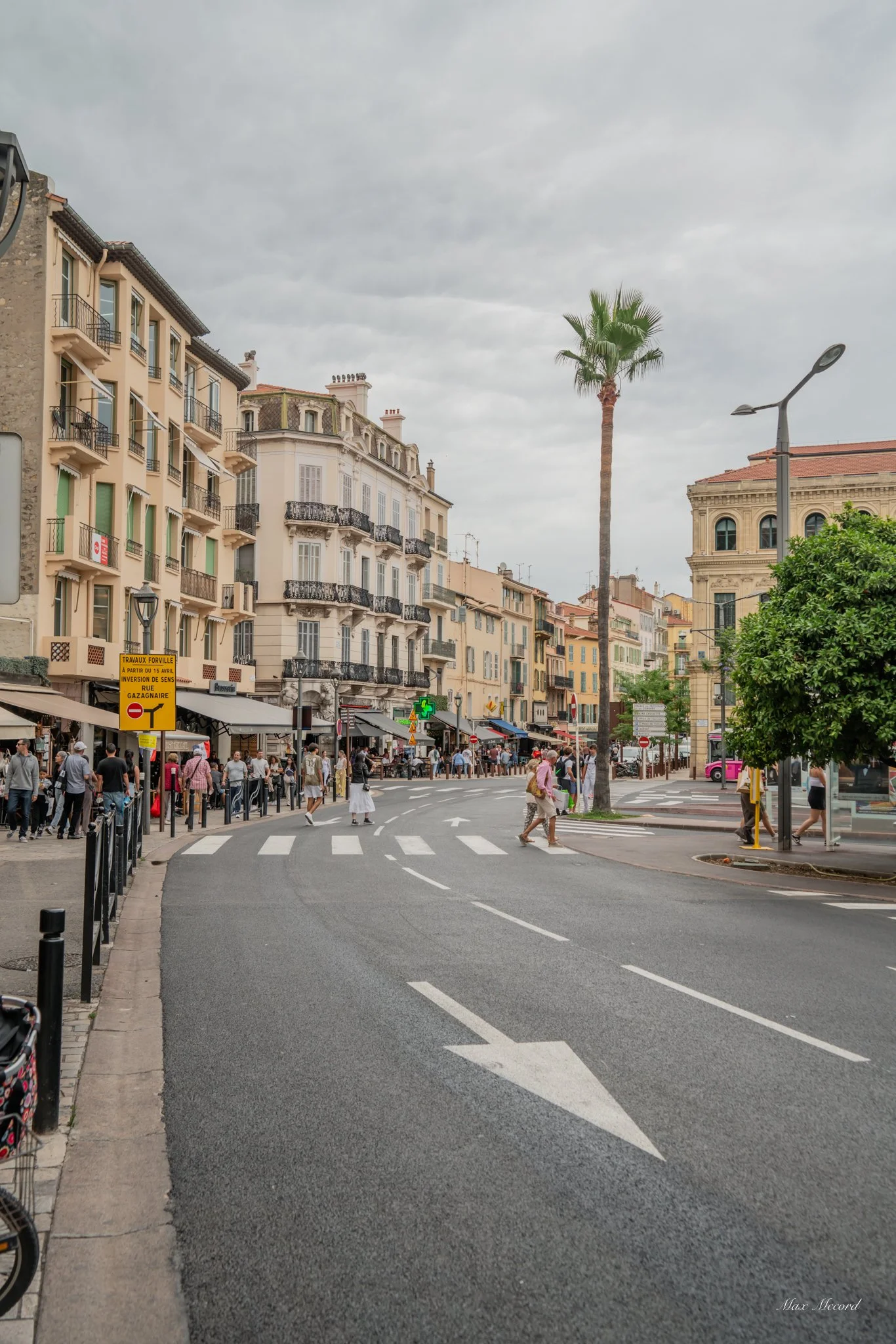 A city street scene with multi-story buildings with balconies, shops, and restaurants on the ground floor. People are crossing the street and walking along the sidewalks. There is a tall palm tree, street signs, and a cloudy sky overhead.