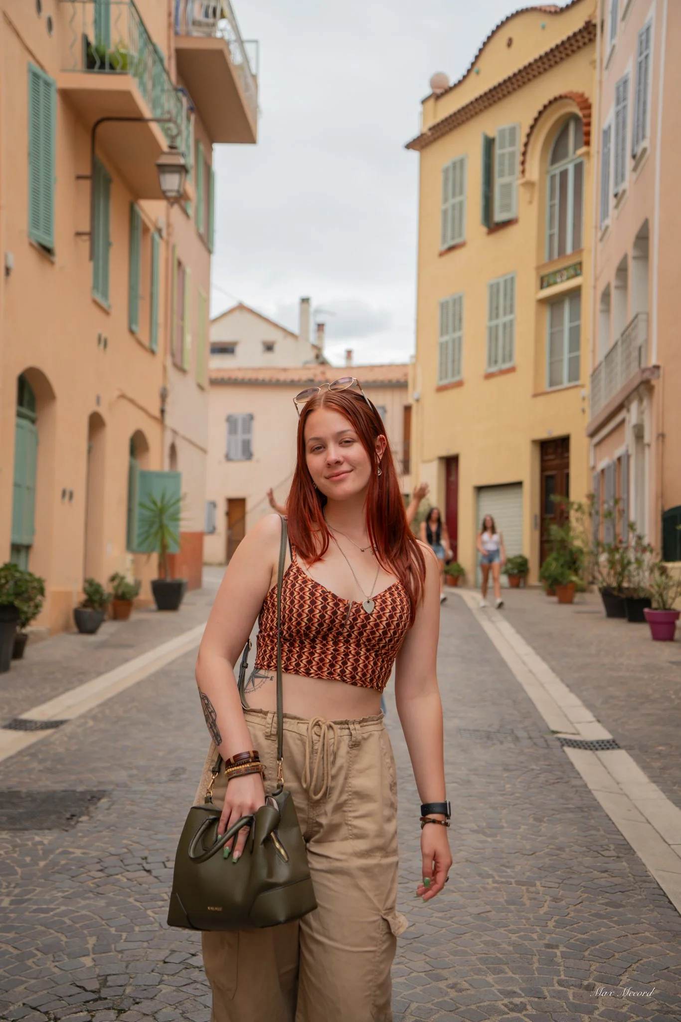 A young woman with red hair standing on a cobblestone street lined with pastel-colored buildings, posing with a slight smile and carrying a black handbag.
