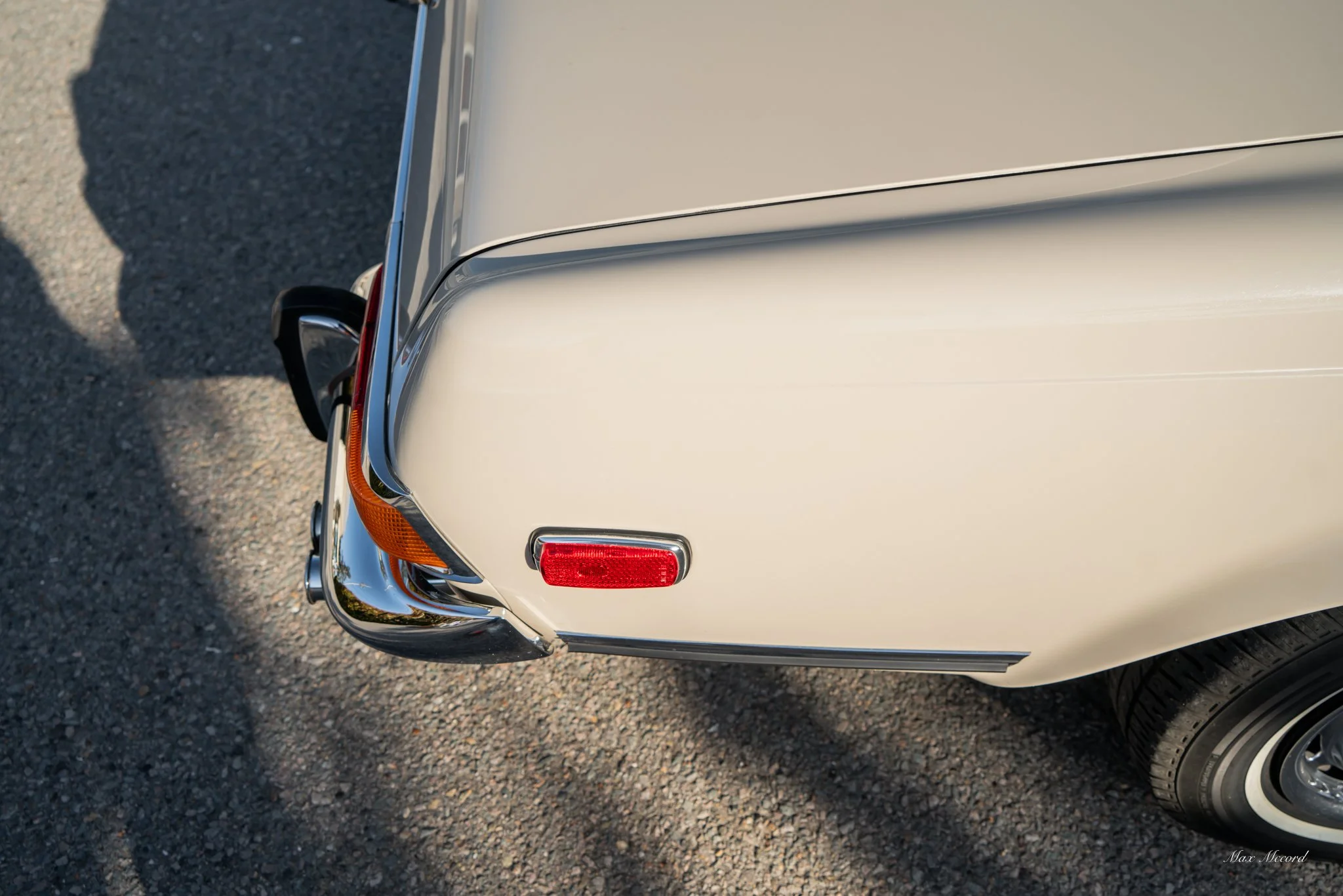 Close-up of the rear corner of a vintage cream-colored car with a chrome bumper, a red reflector, and a round taillight, parked on a gravel surface.