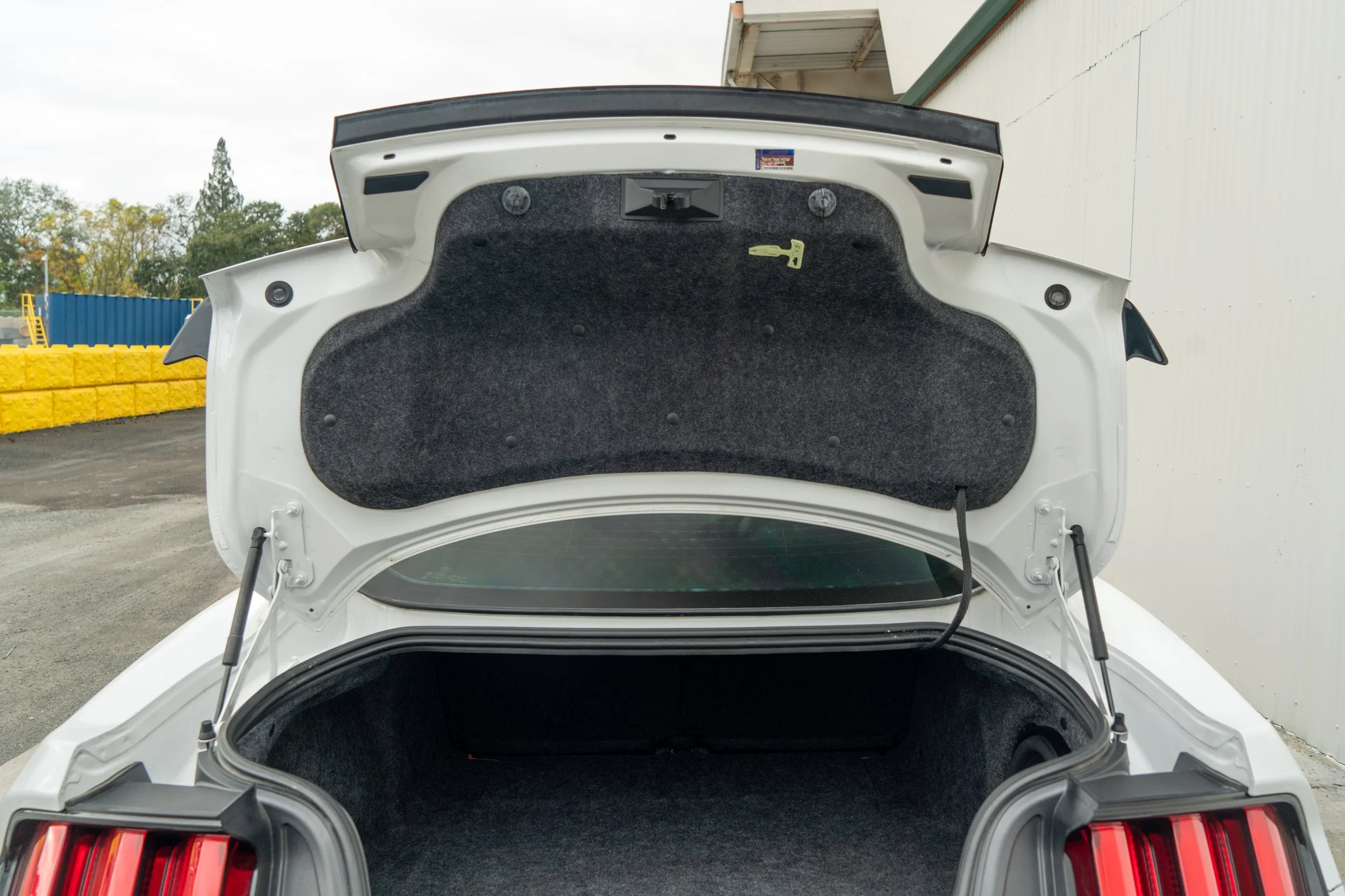 Open trunk of a white car showing gray interior lining and trunk release tools, outdoors near a white building.
