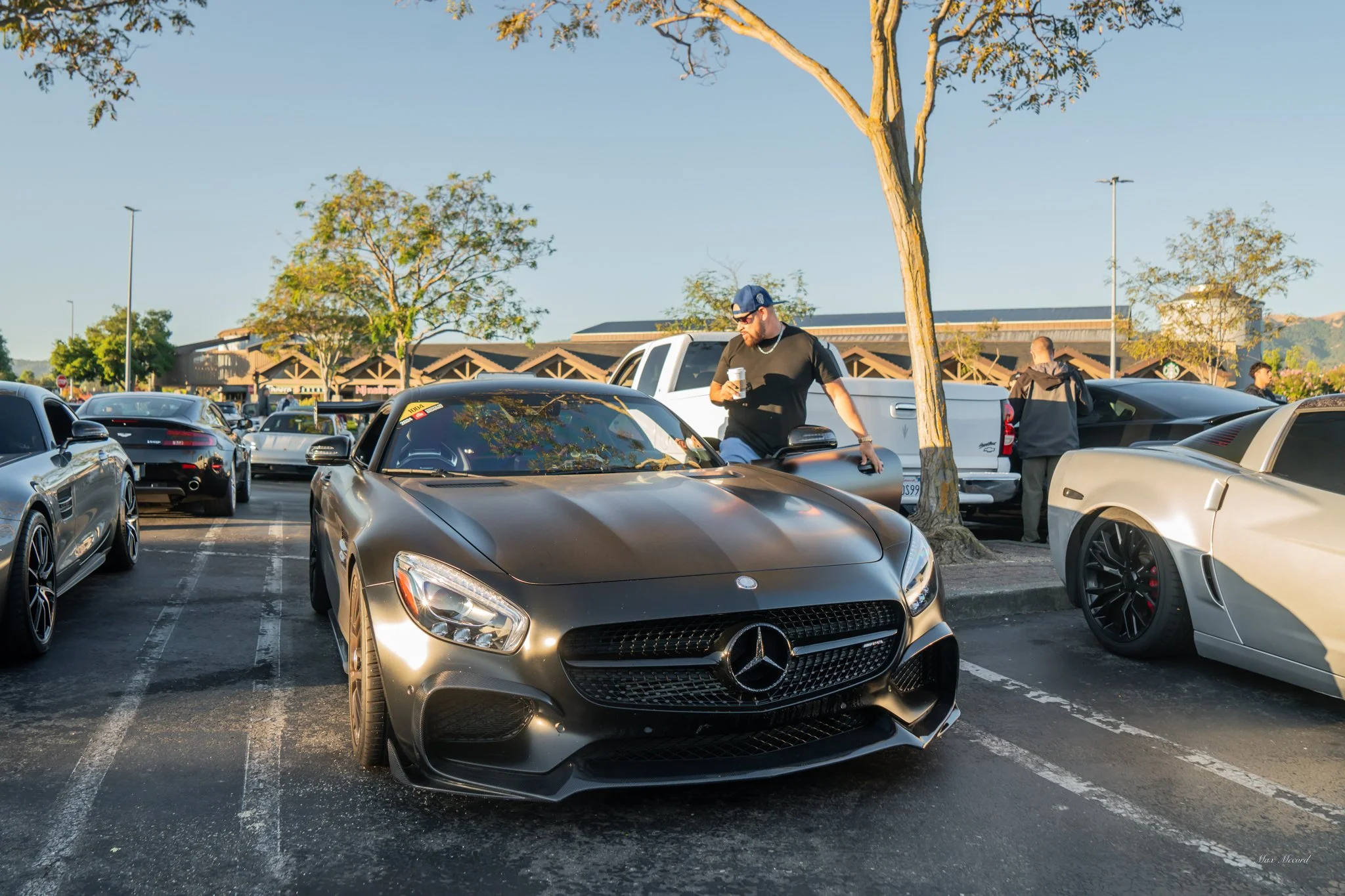 Black Mercedes-Benz sports car parked in a parking lot with a man standing next to it holding a coffee and looking at his phone. Other cars and trees are visible in the background.