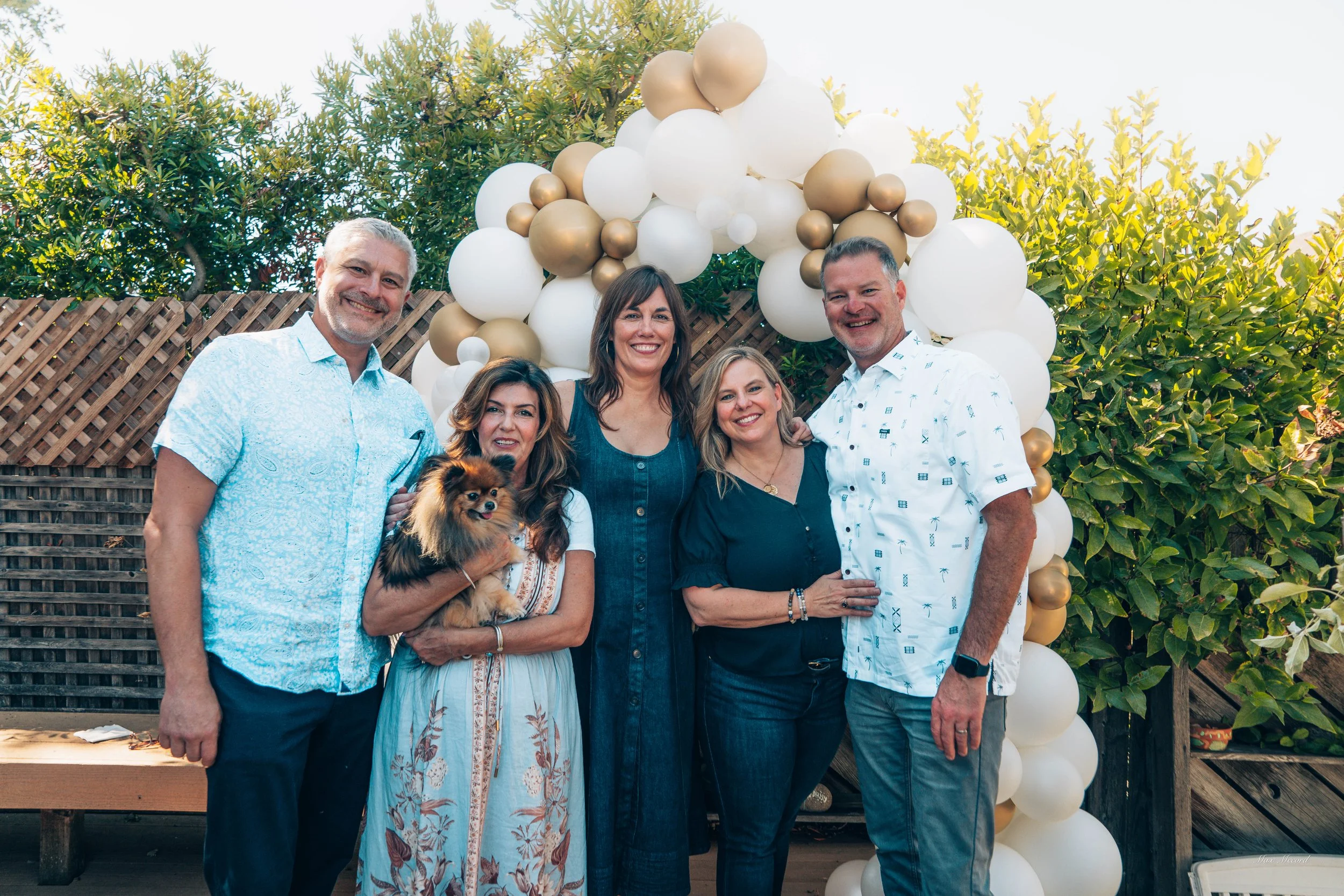 Group of five adults and a small dog standing outdoors in front of a balloon arch and greenery, smiling for the camera.