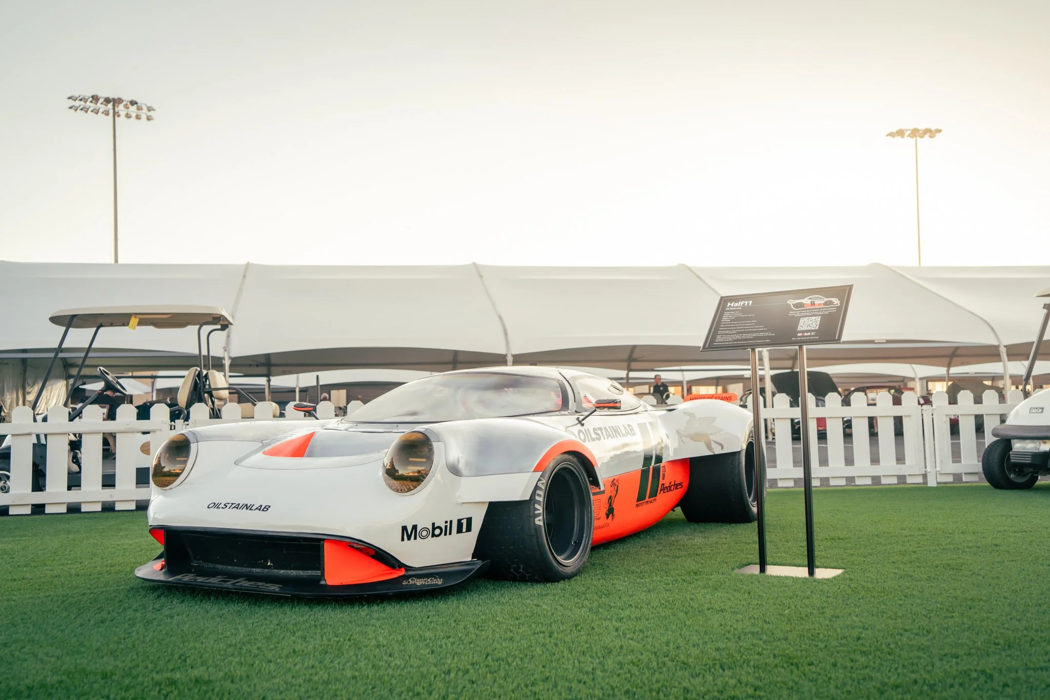 A vintage race car on display at an outdoor event, placed on green artificial grass with a white picket fence and tents in the background.